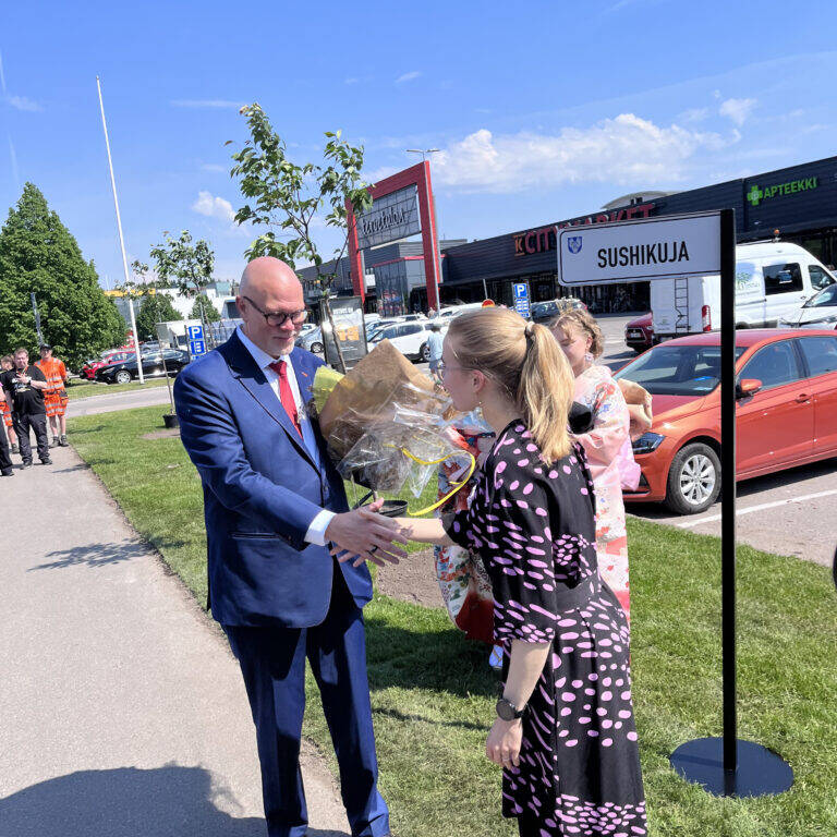 Henkilö ojentaa kukkapuskan puku päällä olevalle henkilölle ja kättelee häntä..