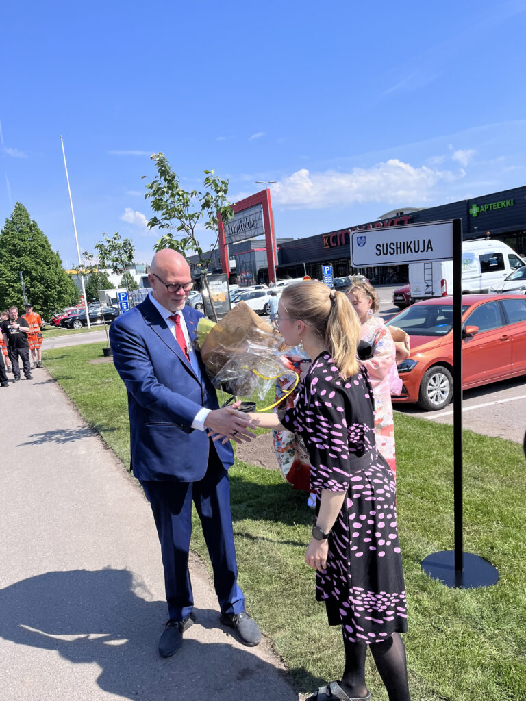 Henkilö ojentaa kukkapuskan puku päällä olevalle henkilölle ja kättelee häntä..