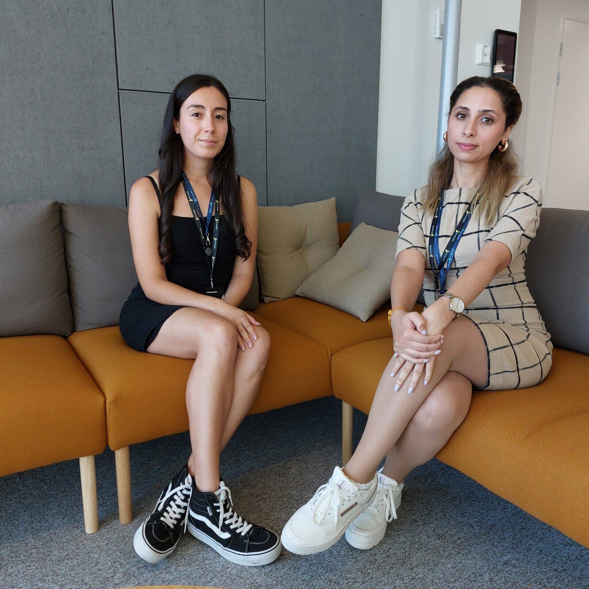 Kaksi naista istuu oranssilla sohvalla modernissa toimistoympäristössä. Vasemmalla oleva nainen on tummatukkainen ja pukeutunut mustaan mekkoon ja mustiin korkeavartisiin tennareihin. Oikealla oleva nainen on vaaleanruskeahiuksinen, pukeutunut beigeen ruudulliseen mekkoon ja valkoisiin platform-tennareihin. Molemmilla on kaulassaan henkilökortit, ja he istuvat jalat ristissä katsoen kameraan.