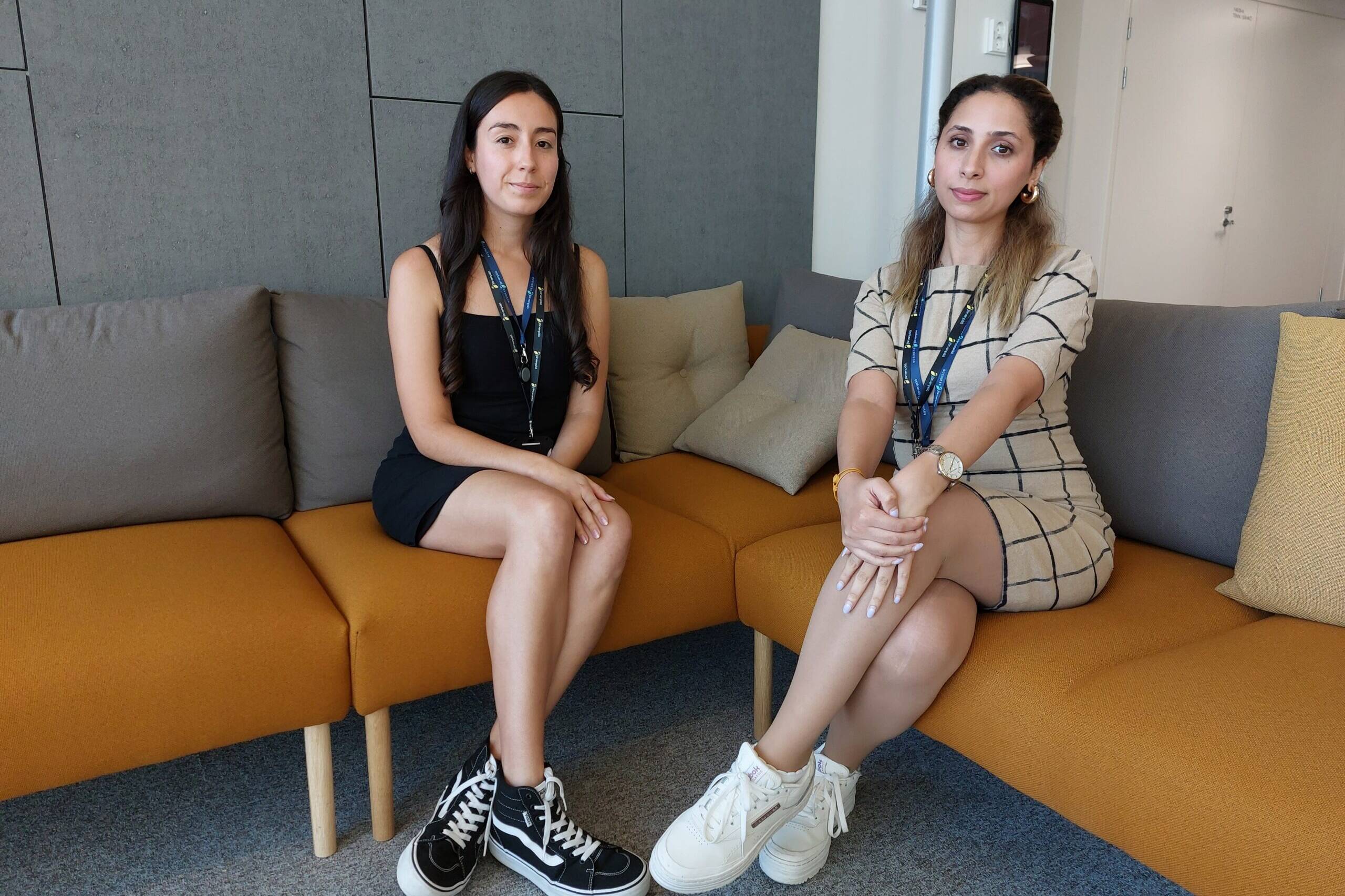 Kaksi naista istuu oranssilla sohvalla modernissa toimistoympäristössä. Vasemmalla oleva nainen on tummatukkainen ja pukeutunut mustaan mekkoon ja mustiin korkeavartisiin tennareihin. Oikealla oleva nainen on vaaleanruskeahiuksinen, pukeutunut beigeen ruudulliseen mekkoon ja valkoisiin platform-tennareihin. Molemmilla on kaulassaan henkilökortit, ja he istuvat jalat ristissä katsoen kameraan.