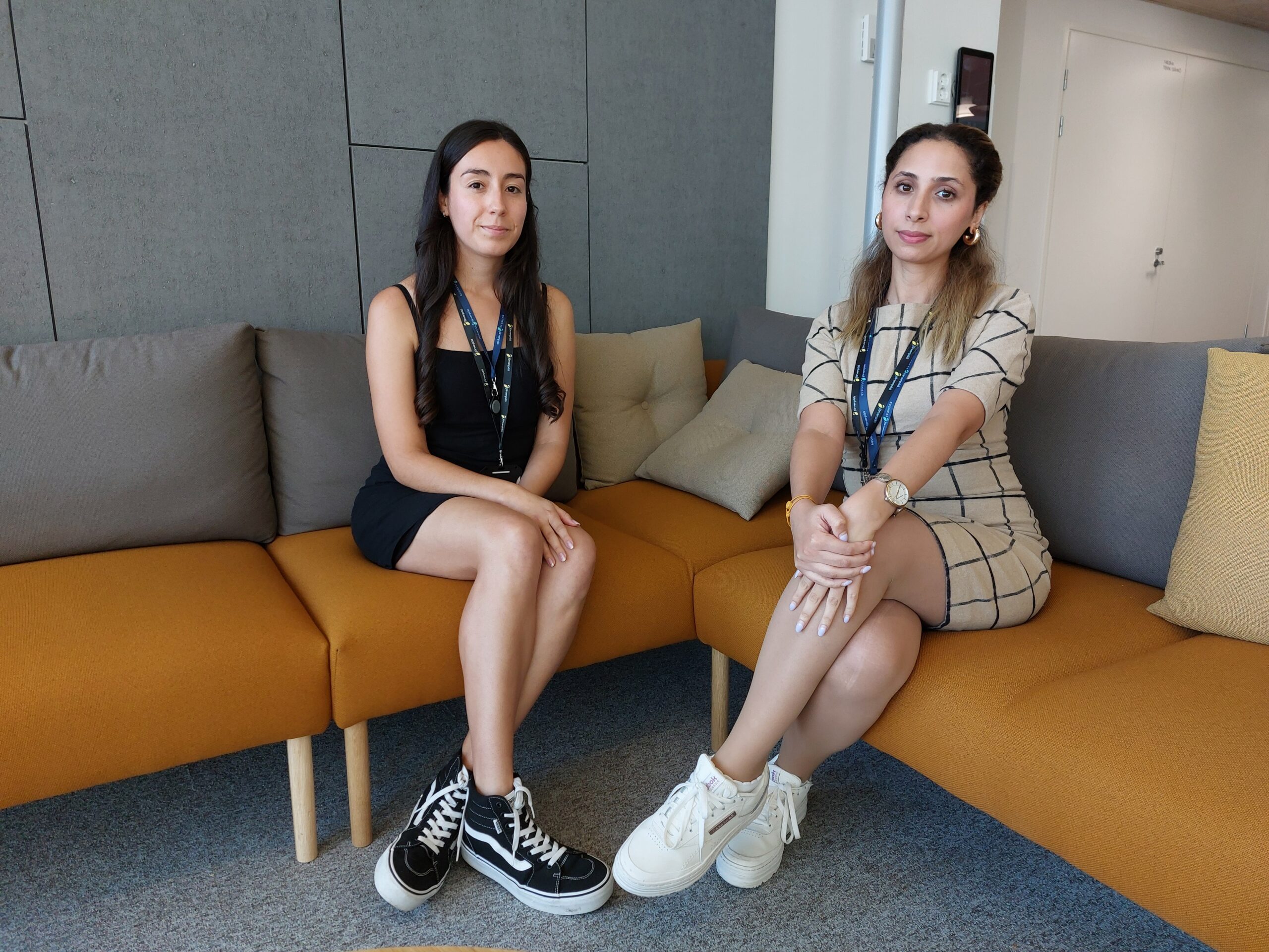 Kaksi naista istuu oranssilla sohvalla modernissa toimistoympäristössä. Vasemmalla oleva nainen on tummatukkainen ja pukeutunut mustaan mekkoon ja mustiin korkeavartisiin tennareihin. Oikealla oleva nainen on vaaleanruskeahiuksinen, pukeutunut beigeen ruudulliseen mekkoon ja valkoisiin platform-tennareihin. Molemmilla on kaulassaan henkilökortit, ja he istuvat jalat ristissä katsoen kameraan.
