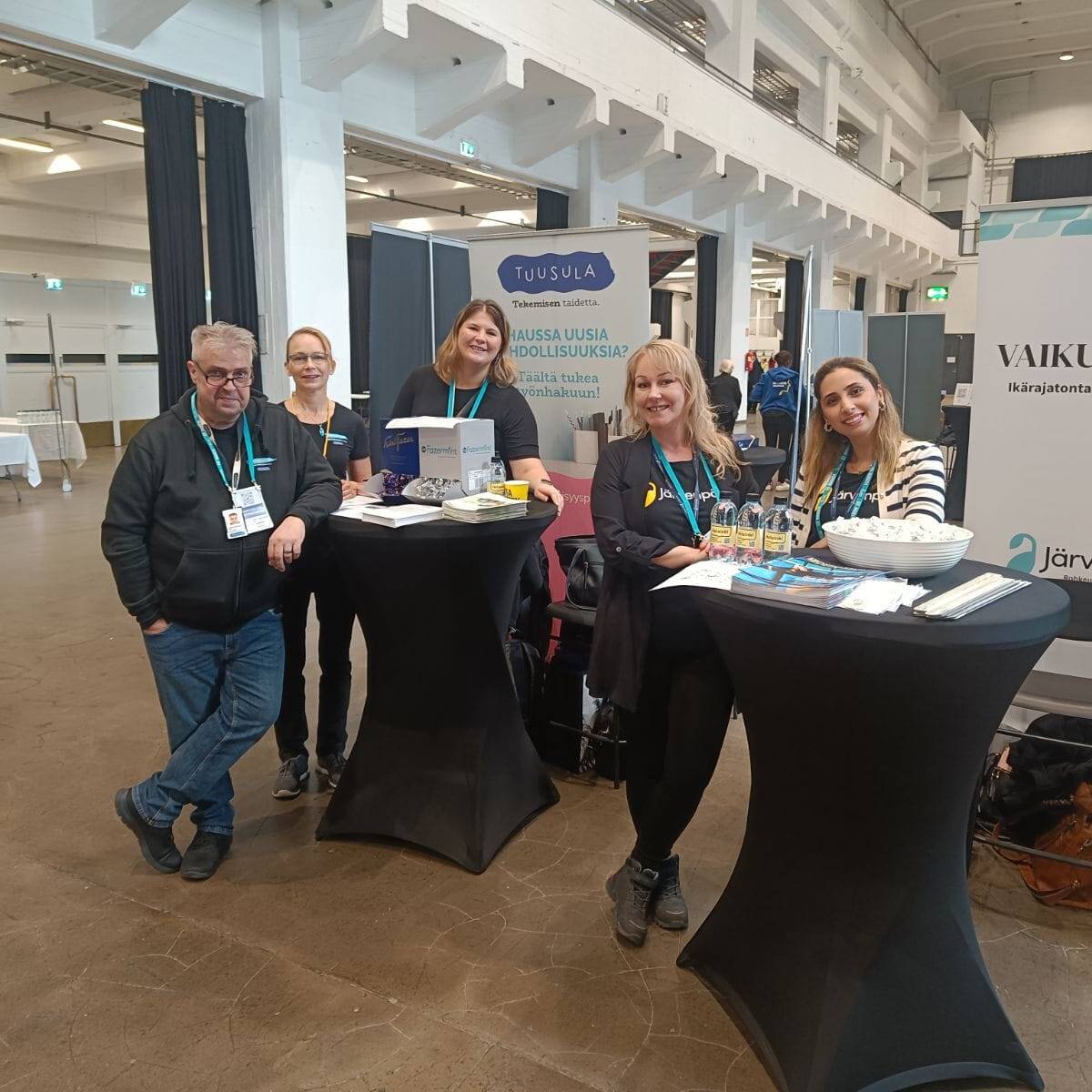 Ryhmä ihmisiä seisoo ja istuu esittelypöytien äärellä Rekrytori for Internationals -tapahtumassa Helsingin Kaapelitehtaalla. Taustalla näkyy Järvenpään ja Tuusulan esittelyosastojen kyltit. Pöydillä on esitteitä ja muita markkinointimateriaaleja, ja osallistujat hymyilevät kameralle.