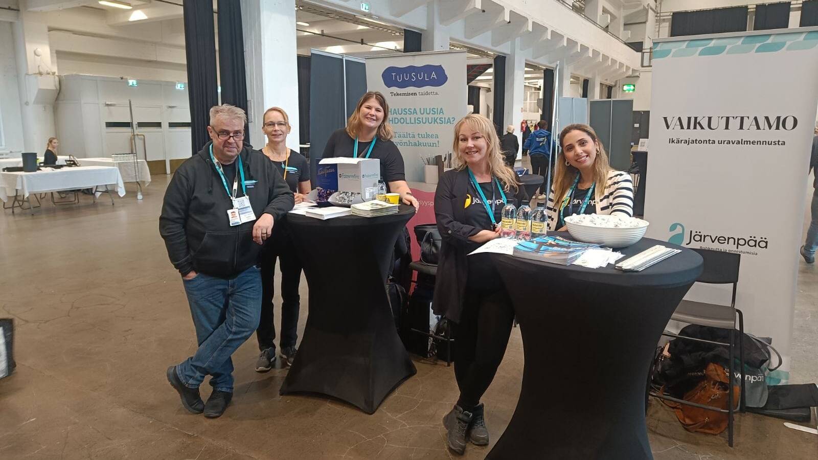 Ryhmä ihmisiä seisoo ja istuu esittelypöytien äärellä Rekrytori for Internationals -tapahtumassa Helsingin Kaapelitehtaalla. Taustalla näkyy Järvenpään ja Tuusulan esittelyosastojen kyltit. Pöydillä on esitteitä ja muita markkinointimateriaaleja, ja osallistujat hymyilevät kameralle.