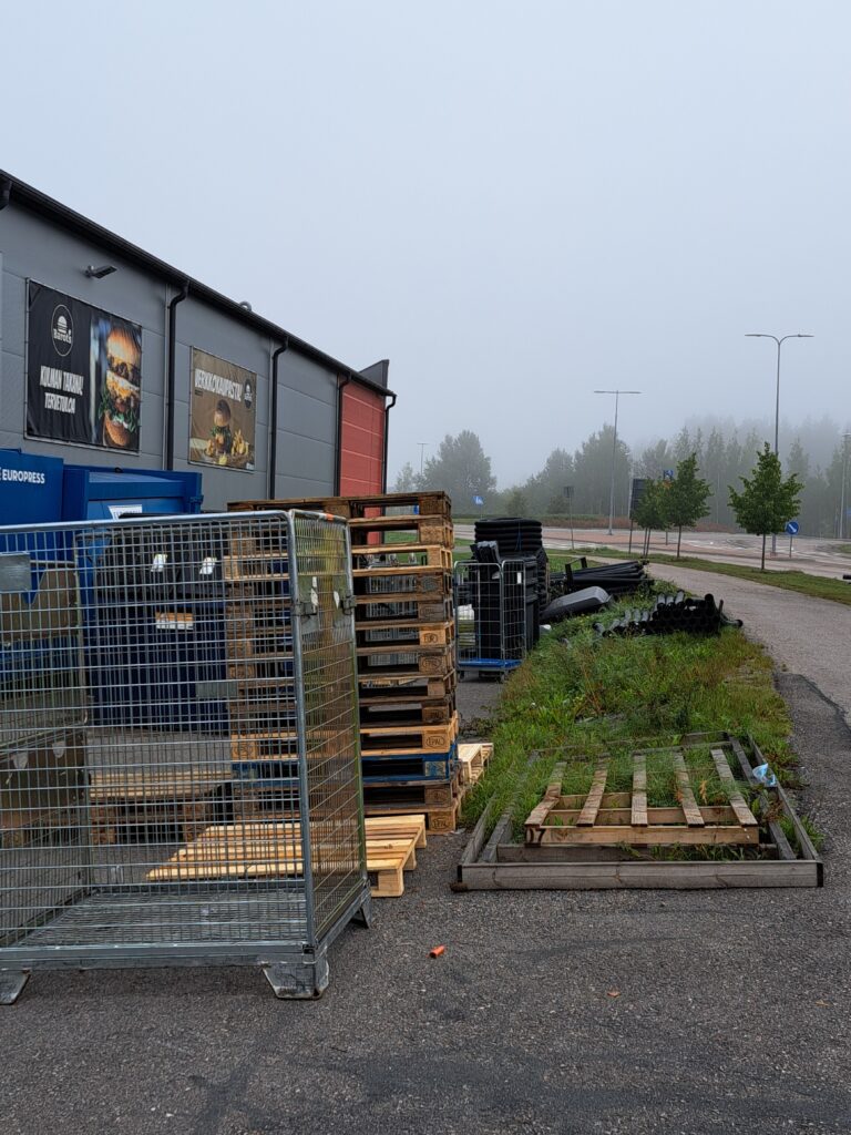 Sumuinen näkymä varaston tai teollisuusrakennuksen pihalta. Kuvassa on puisia lavoja, metallihäkkejä ja rakennusmateriaaleja. Rakennuksen seinällä on mainoskyltit, joissa näkyy hampurilaisia. Oikealla on tie ja puita.