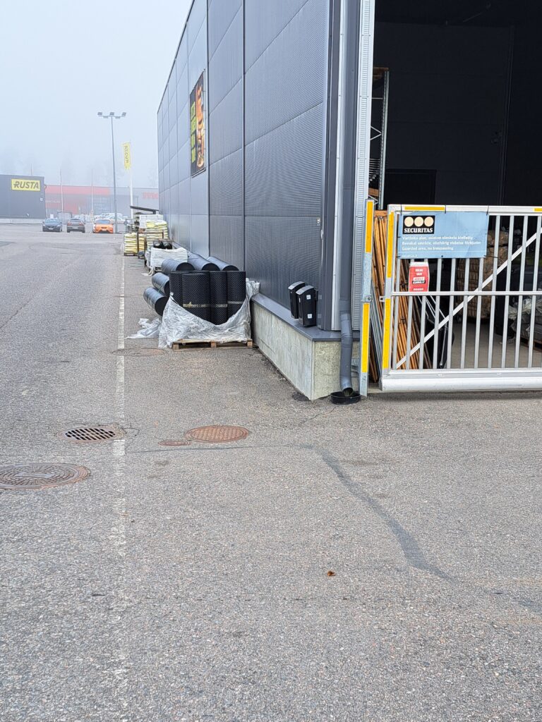 Sumuinen näkymä varastorakennuksen sivulta. Kuvassa on muoviputkia ja rakennustarvikkeita lavoilla seinän vieressä. Rakennuksessa näkyy Securitaksen varoituskyltti portin vieressä, ja taustalla on kauppojen kylttejä, kuten Rusta.