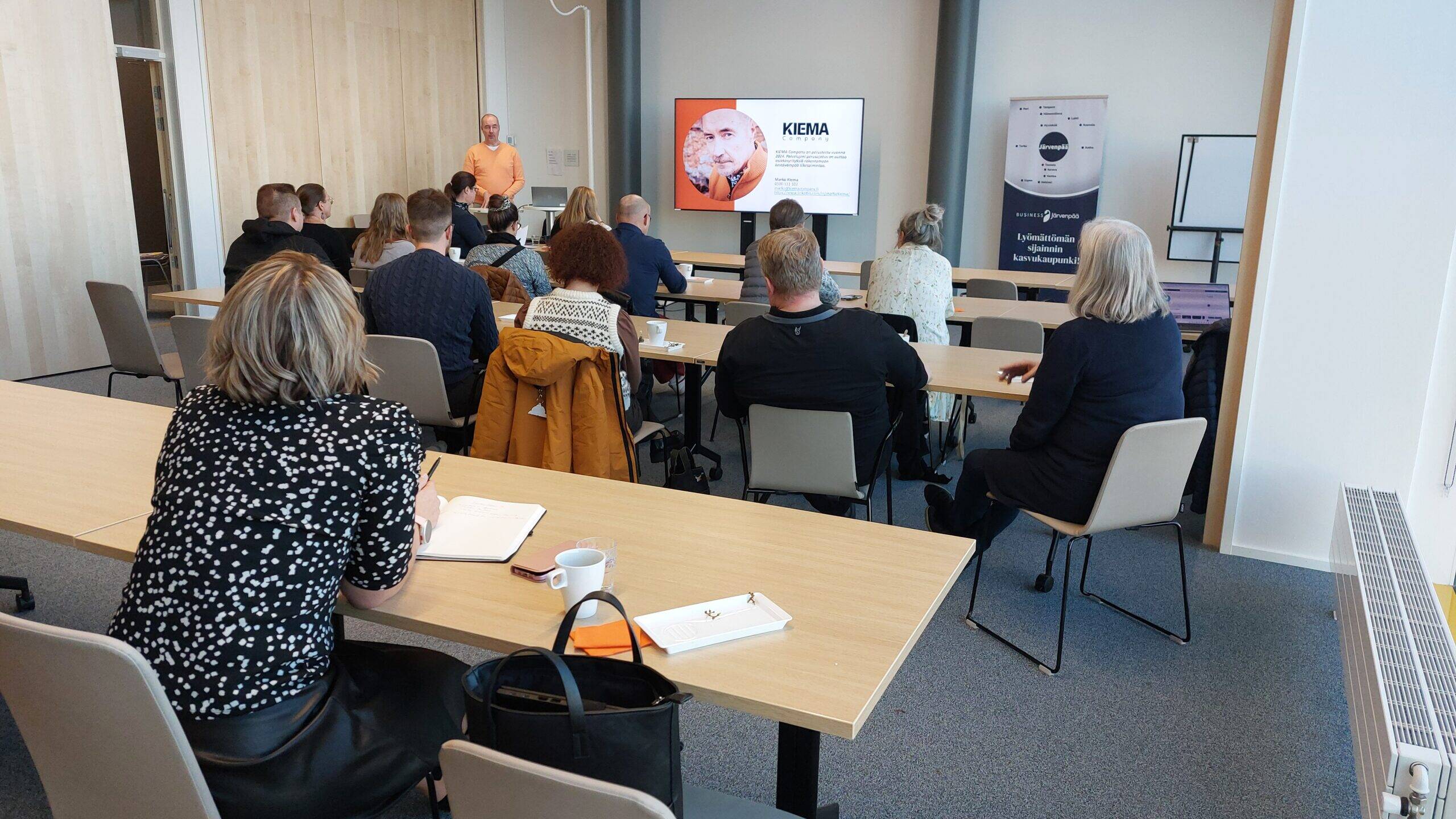 Henkilö puhumassa yritysyleisölle kokoustilassa, jossa on puinen seinäpaneeli ja suuret ikkunat. Seinällä näkyy valkokangas, jossa lukee 