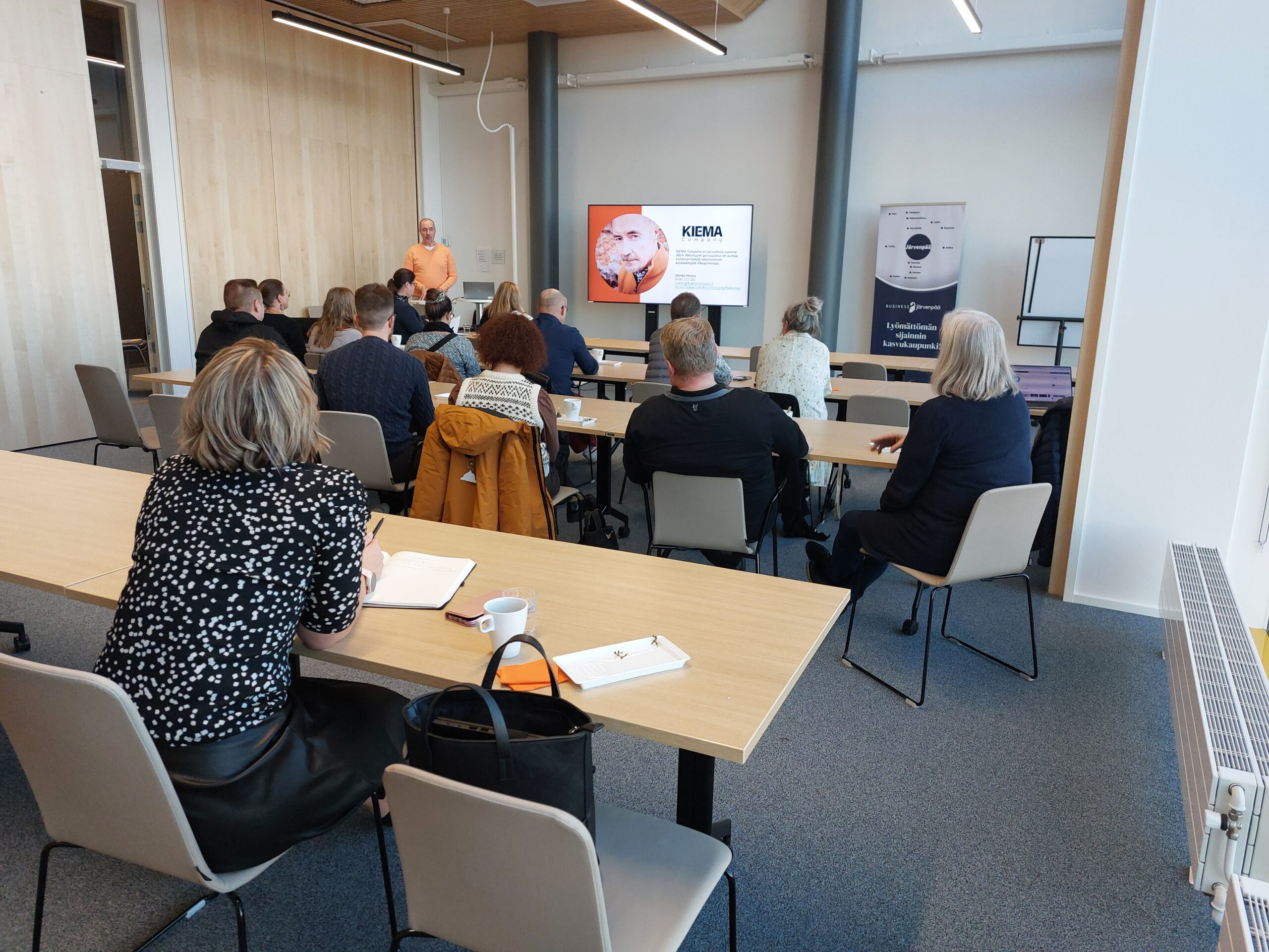 Henkilö puhumassa yritysyleisölle kokoustilassa, jossa on puinen seinäpaneeli ja suuret ikkunat. Seinällä näkyy valkokangas, jossa lukee 