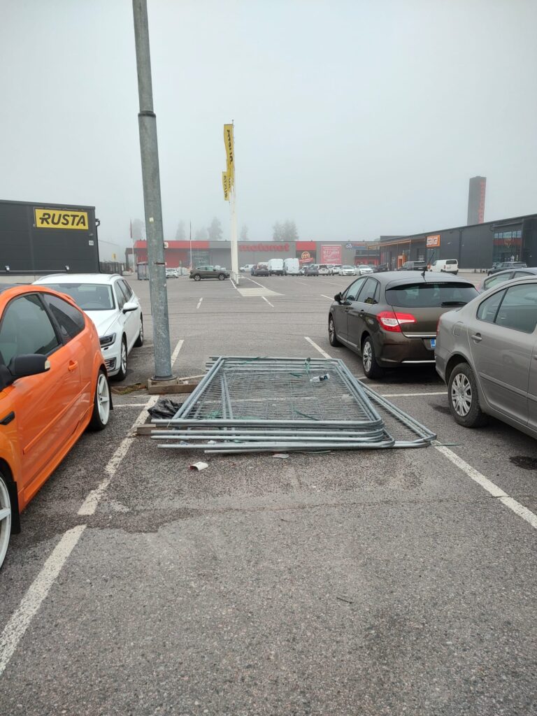 Kuvassa on parkkipaikka, jossa on autoja pysäköitynä, ja maassa on metallisia aitaelementtejä kasassa. Taustalla näkyy kauppakeskus, jossa on liikkeitä, kuten Rusta ja Motonet, ja muutama kaupan logo on näkyvissä. Sumuinen sää luo pehmeän ja hieman utuinen tunnelman.