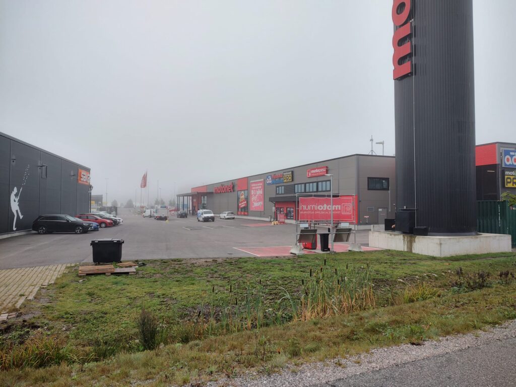 Kuvassa on kauppakeskusalue, jossa on liikkeitä, kuten Motonet ja Gigantti. Parkkipaikalla on muutamia autoja, ja sää on sumuinen, mikä pehmentää taustan yksityiskohtia. Vasemmalla näkyy suuri seinämaalaus Gigantin rakennuksessa, ja oikealla on Motonetin korkea kyltti. Etualalla on vihreää nurmialuetta, jossa on matalaa kasvillisuutta.