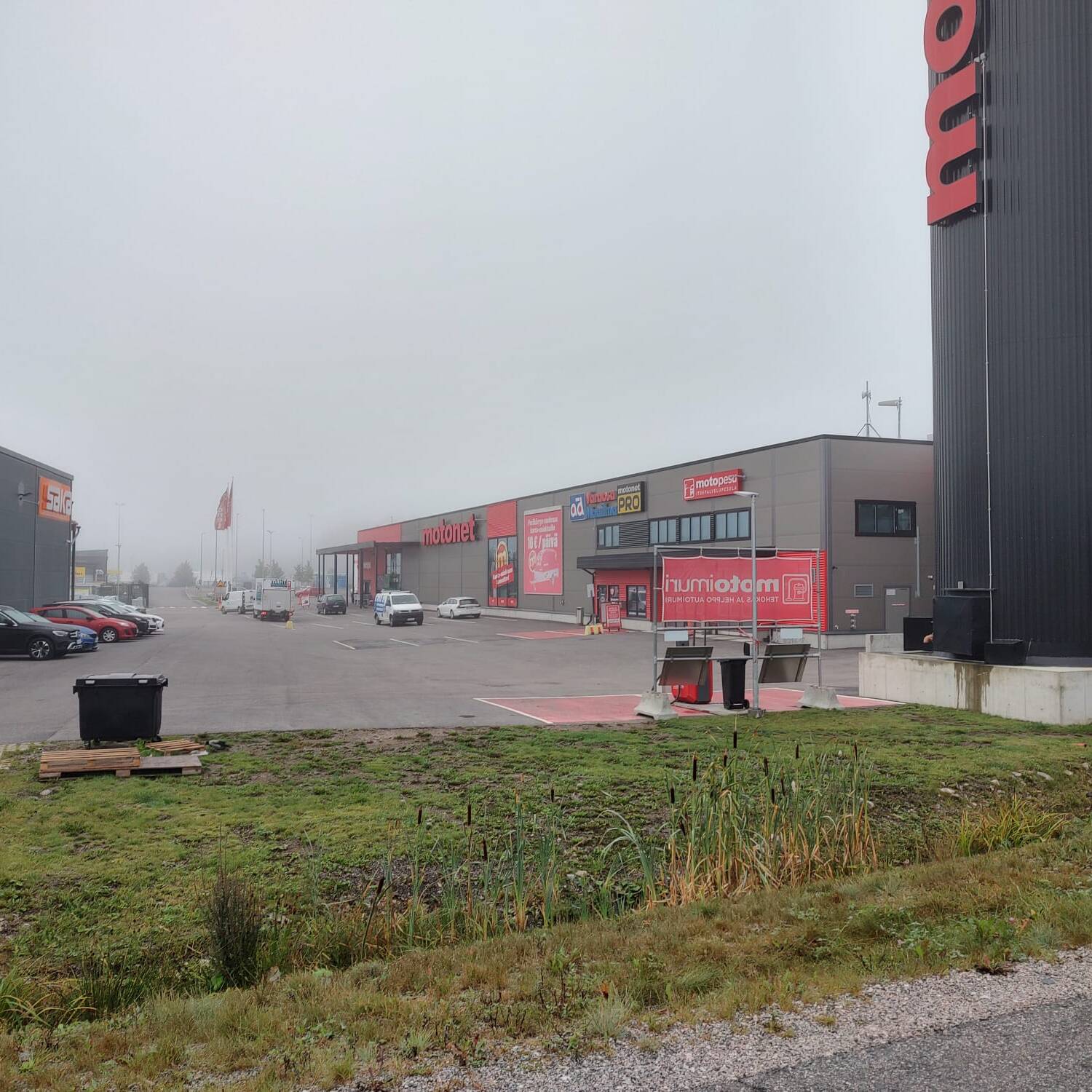 Kuvassa on kauppakeskusalue, jossa on liikkeitä, kuten Motonet ja Gigantti. Parkkipaikalla on muutamia autoja, ja sää on sumuinen, mikä pehmentää taustan yksityiskohtia. Vasemmalla näkyy suuri seinämaalaus Gigantin rakennuksessa, ja oikealla on Motonetin korkea kyltti. Etualalla on vihreää nurmialuetta, jossa on matalaa kasvillisuutta.