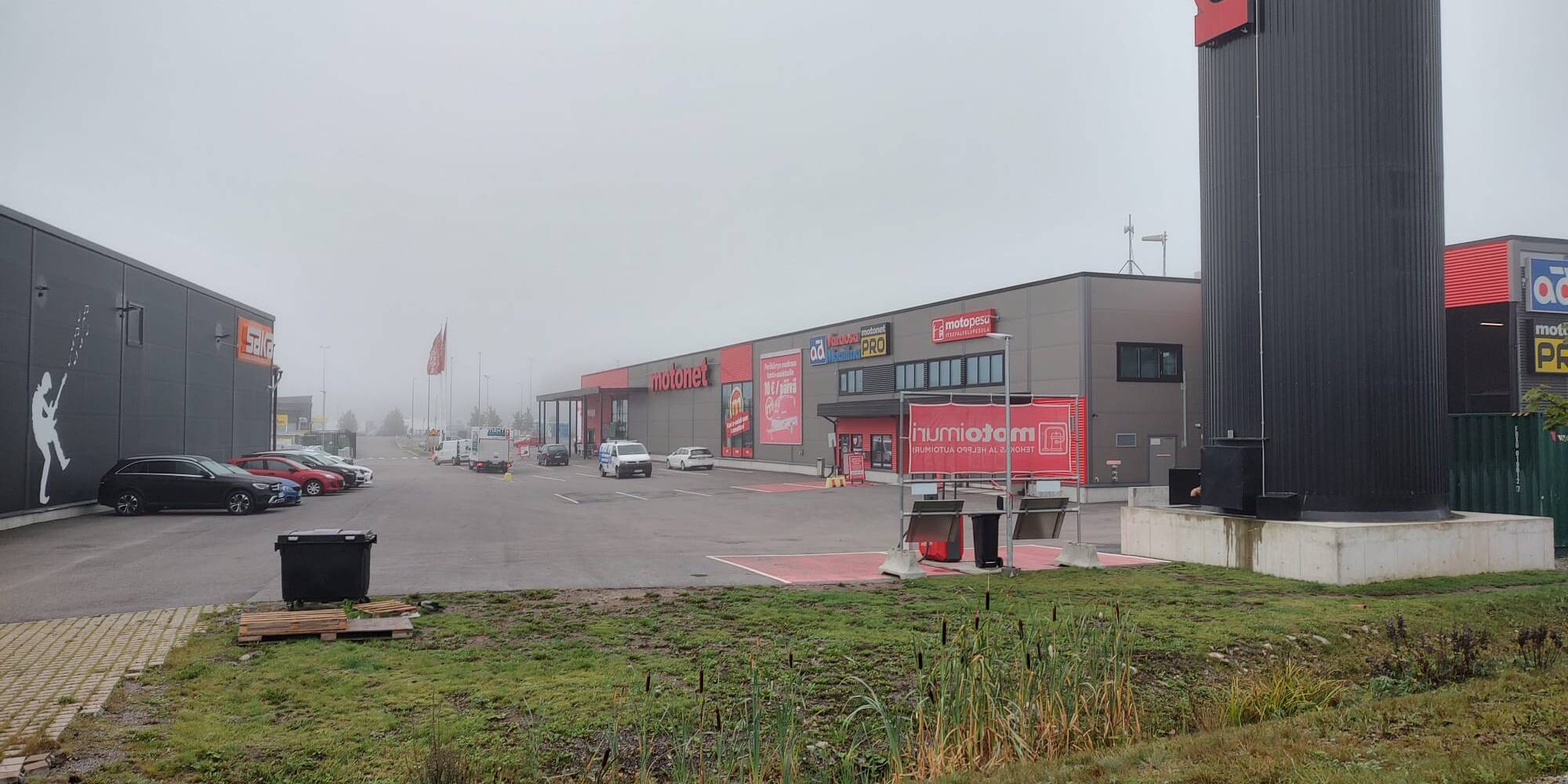 Kuvassa on kauppakeskusalue, jossa on liikkeitä, kuten Motonet ja Gigantti. Parkkipaikalla on muutamia autoja, ja sää on sumuinen, mikä pehmentää taustan yksityiskohtia. Vasemmalla näkyy suuri seinämaalaus Gigantin rakennuksessa, ja oikealla on Motonetin korkea kyltti. Etualalla on vihreää nurmialuetta, jossa on matalaa kasvillisuutta.