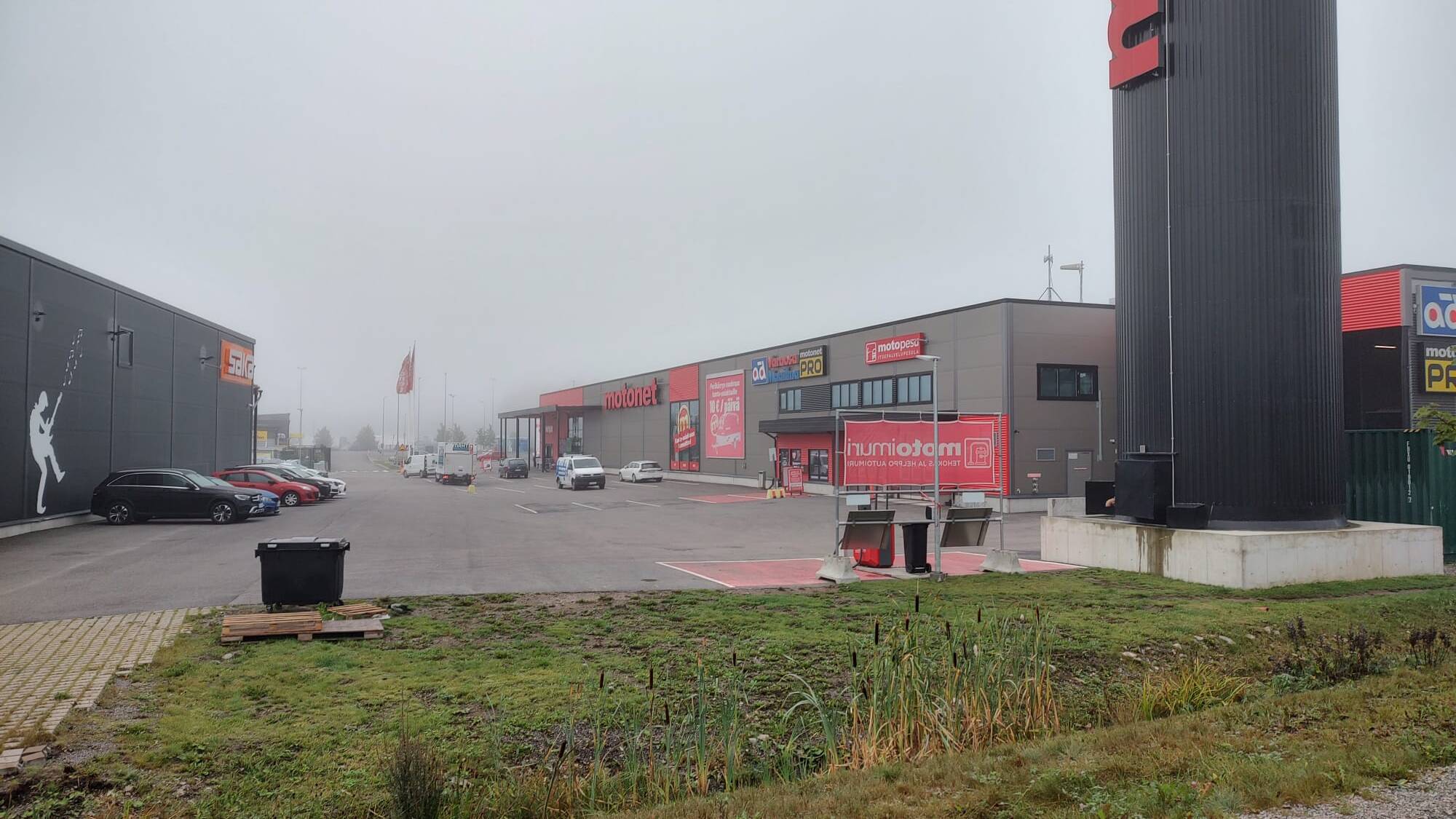 Kuvassa on kauppakeskusalue, jossa on liikkeitä, kuten Motonet ja Gigantti. Parkkipaikalla on muutamia autoja, ja sää on sumuinen, mikä pehmentää taustan yksityiskohtia. Vasemmalla näkyy suuri seinämaalaus Gigantin rakennuksessa, ja oikealla on Motonetin korkea kyltti. Etualalla on vihreää nurmialuetta, jossa on matalaa kasvillisuutta.