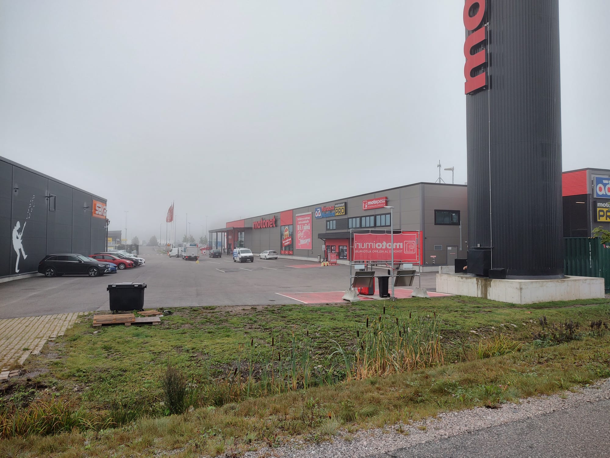 Kuvassa on kauppakeskusalue, jossa on liikkeitä, kuten Motonet ja Gigantti. Parkkipaikalla on muutamia autoja, ja sää on sumuinen, mikä pehmentää taustan yksityiskohtia. Vasemmalla näkyy suuri seinämaalaus Gigantin rakennuksessa, ja oikealla on Motonetin korkea kyltti. Etualalla on vihreää nurmialuetta, jossa on matalaa kasvillisuutta.