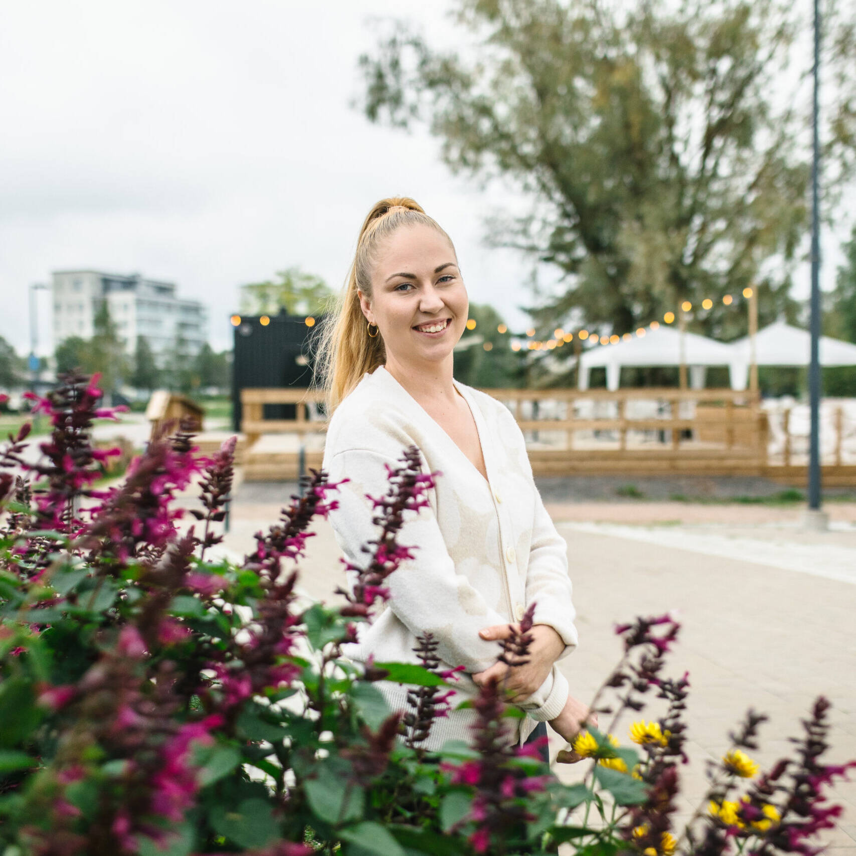 Kuvassa näkyy nainen, joka seisoo puistomaisessa ympäristössä kukkien vieressä. Hän hymyilee kameralle ja vaikuttaa rennolta. Taustalla on ulkoilma-alue, jossa on puinen terassi ja valaistus, ehkä valonauhoja, jotka tuovat tunnelmaa alueelle. Myös puita ja pensaita sekä moderneja rakennuksia näkyy taustalla. Tämä ympäristö näyttää olevan osa tapahtuma- tai kahvilapaikkaa, mahdollisesti samaa 