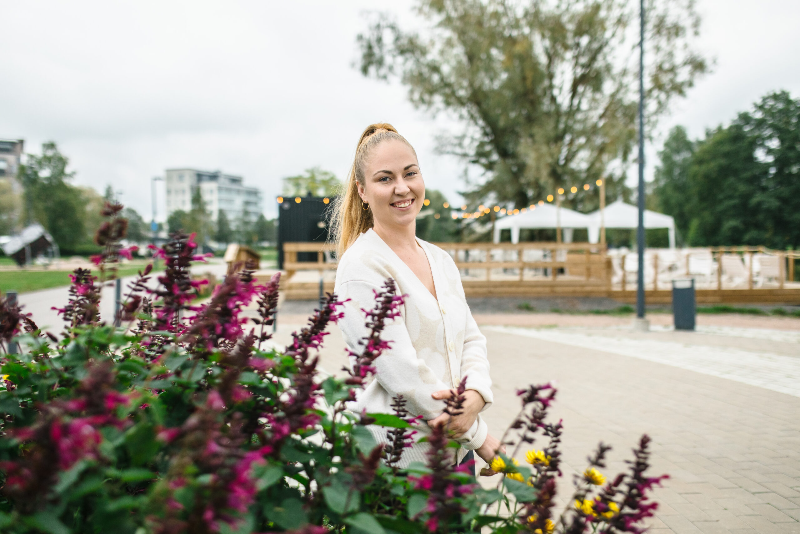 Kuvassa näkyy nainen, joka seisoo puistomaisessa ympäristössä kukkien vieressä. Hän hymyilee kameralle ja vaikuttaa rennolta. Taustalla on ulkoilma-alue, jossa on puinen terassi ja valaistus, ehkä valonauhoja, jotka tuovat tunnelmaa alueelle. Myös puita ja pensaita sekä moderneja rakennuksia näkyy taustalla. Tämä ympäristö näyttää olevan osa tapahtuma- tai kahvilapaikkaa, mahdollisesti samaa "AETO Garden" -ympäristöä kuin aiemmissa kuvissa. Paikka näyttää viihtyisältä ja ulkoilmatilaan suunnitellulta sosiaalisen kanssakäymisen alueelta.