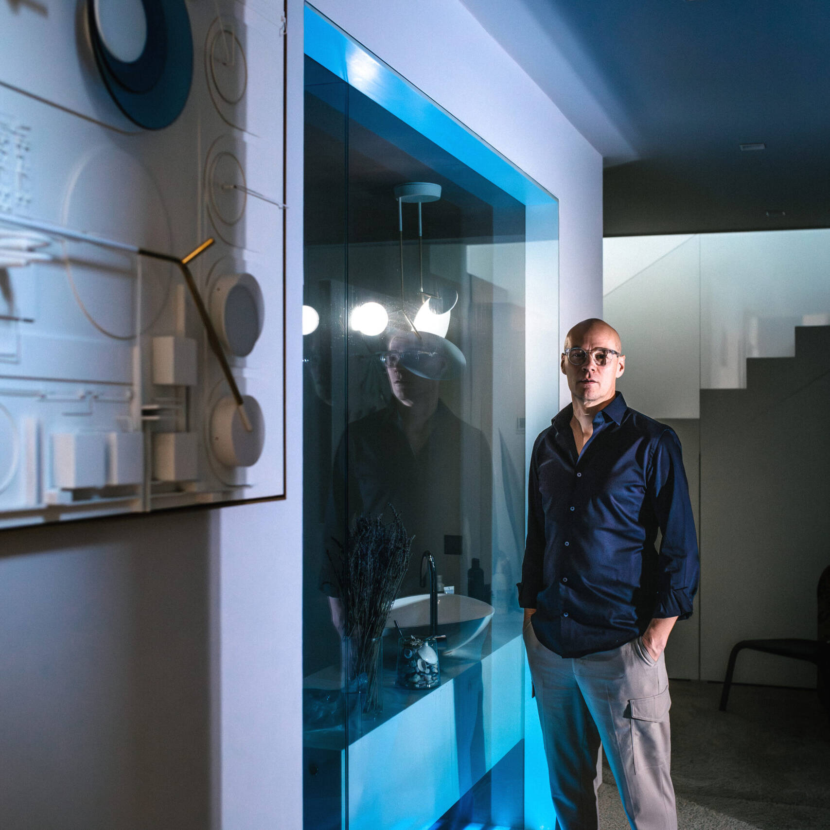 Mies seisoo modernissa sisätilassa valaistun taideteoksen edessä. Seinällä on valkoista ja sinistä sisältävä kohokuvioinen taideteos. Taustalla näkyy lasinen pesuallasalue ja portaikko.