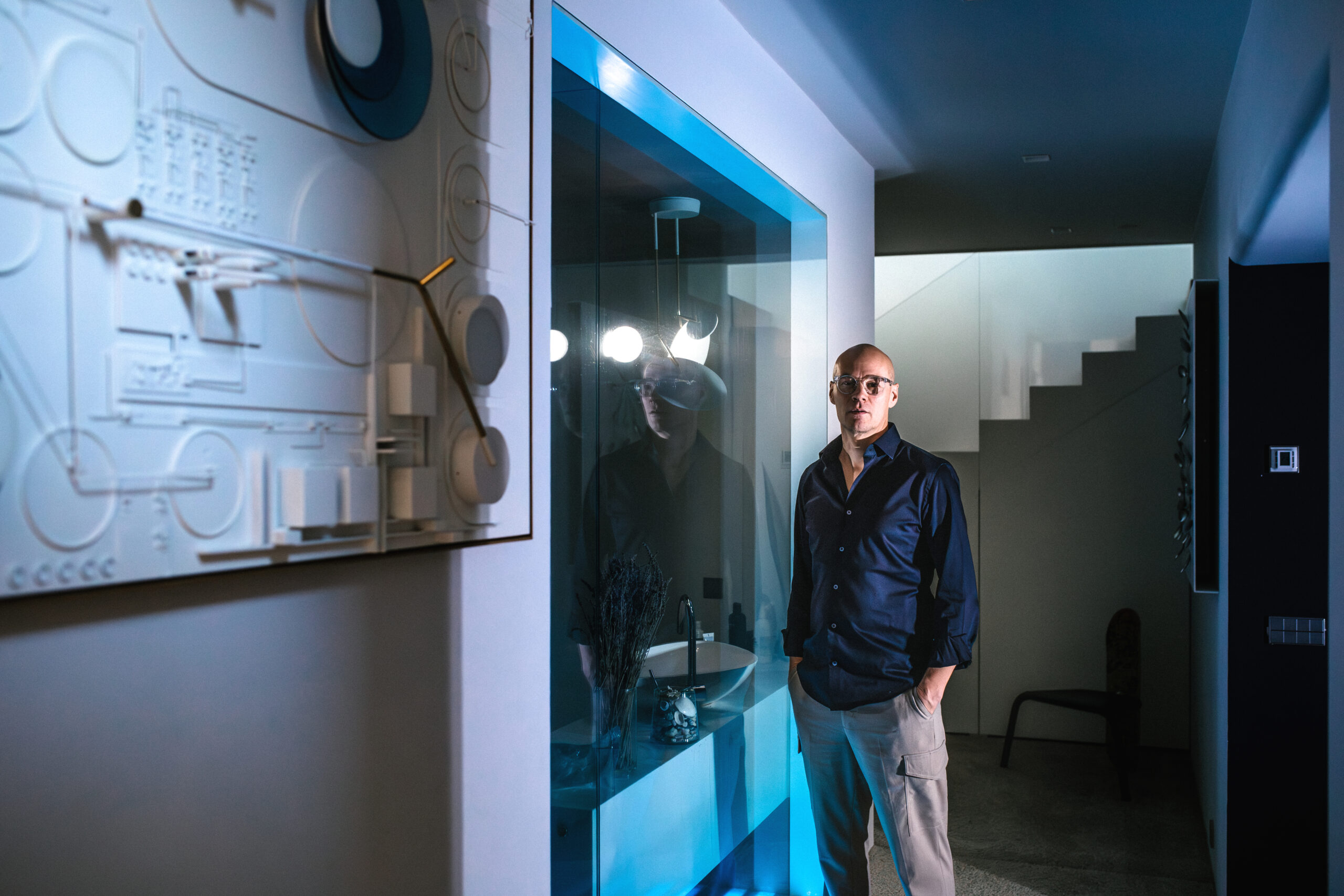 Mies seisoo modernissa sisätilassa valaistun taideteoksen edessä. Seinällä on valkoista ja sinistä sisältävä kohokuvioinen taideteos. Taustalla näkyy lasinen pesuallasalue ja portaikko.