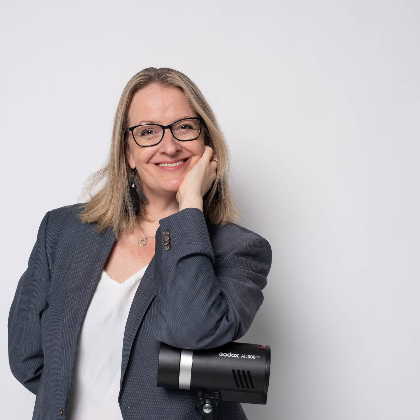 Hymyilevä henkilö seisoo vaaleaa taustaa vasten, pukeutuneena harmaaseen bleiseriin ja valkoiseen puseroon. Hän nojaa kädellään poskeensa ja pitää toisessa kädessään Godox AD300Pro -studiosalamaa.
