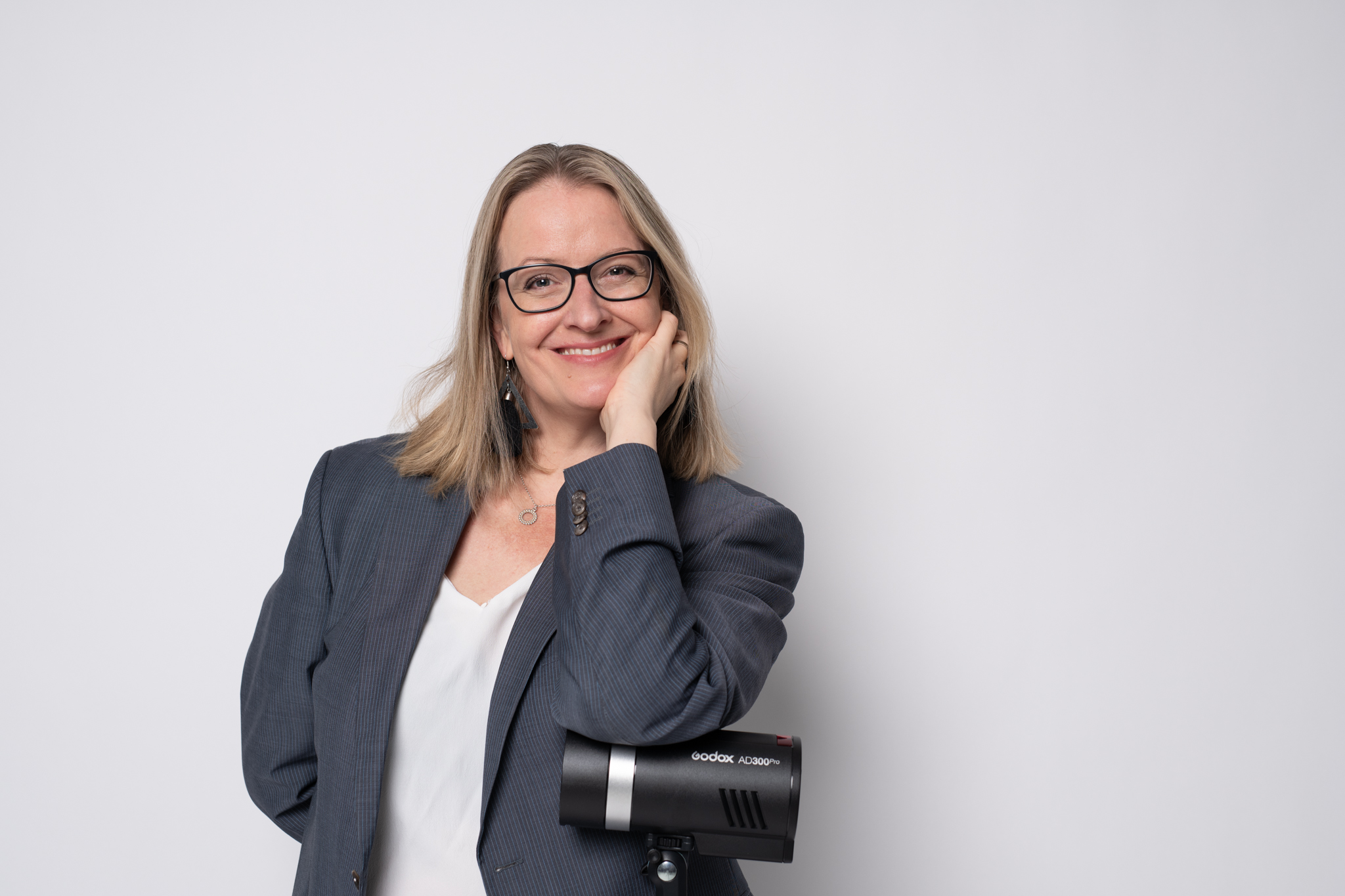 Hymyilevä henkilö seisoo vaaleaa taustaa vasten, pukeutuneena harmaaseen bleiseriin ja valkoiseen puseroon. Hän nojaa kädellään poskeensa ja pitää toisessa kädessään Godox AD300Pro -studiosalamaa.