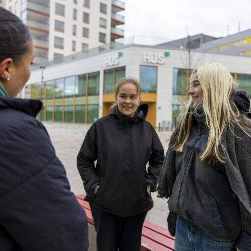 Kolme nuorta naista seisoo ulkona keskustellen keskenään. Taustalla näkyy HUSin rakennus ja moderneja kerrostaloja. Kaikki ovat pukeutuneet lämpimiin ulkovaatteisiin, ja he näyttävät iloisilta ja rennolta.