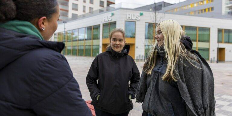 Kolme nuorta naista seisoo ulkona keskustellen keskenään. Taustalla näkyy HUSin rakennus ja moderneja kerrostaloja. Kaikki ovat pukeutuneet lämpimiin ulkovaatteisiin, ja he näyttävät iloisilta ja rennolta.