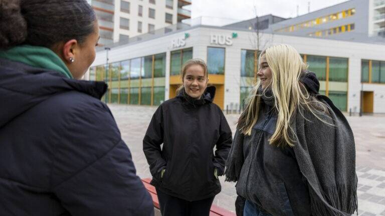 Kolme nuorta naista seisoo ulkona keskustellen keskenään. Taustalla näkyy HUSin rakennus ja moderneja kerrostaloja. Kaikki ovat pukeutuneet lämpimiin ulkovaatteisiin, ja he näyttävät iloisilta ja rennolta.