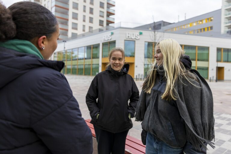 Kolme nuorta naista seisoo ulkona keskustellen keskenään. Taustalla näkyy HUSin rakennus ja moderneja kerrostaloja. Kaikki ovat pukeutuneet lämpimiin ulkovaatteisiin, ja he näyttävät iloisilta ja rennolta.