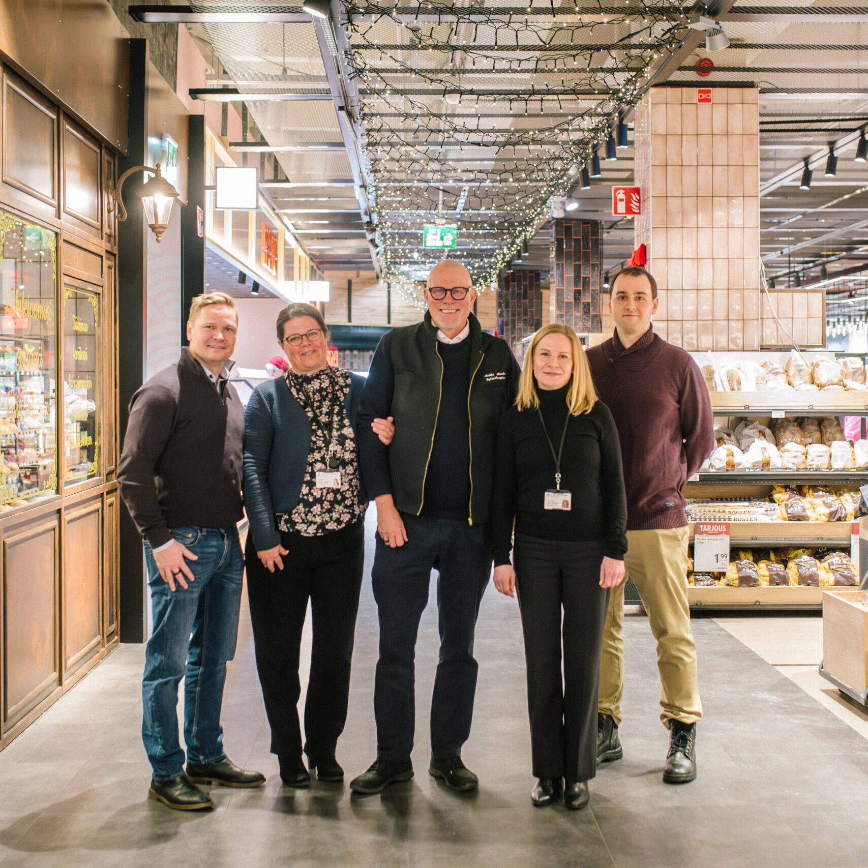 Kuvassa viisi henkilöä seisoo hymyillen ruokakaupan käytävällä. Taustalla näkyy leipäosasto ja myymälän sisustusta, jossa on lämminhenkinen valaistus ja koristevaloja katossa. Vasemmalla puolella on tyylikkäästi somistettu lasivitriini, jossa lukee koristeellisella tekstillä suomenkielisiä sanoja. Oikealla puolella näkyy leipähyllyjä, joissa on tarjouksia ja tuotteita esillä. Kaikilla henkilöillä on siistit vaatteet, ja osalla heistä on nimikortit kaulassaan, viitaten mahdollisesti kaupan henkilökuntaan tai yhteistyökumppaneihin.
