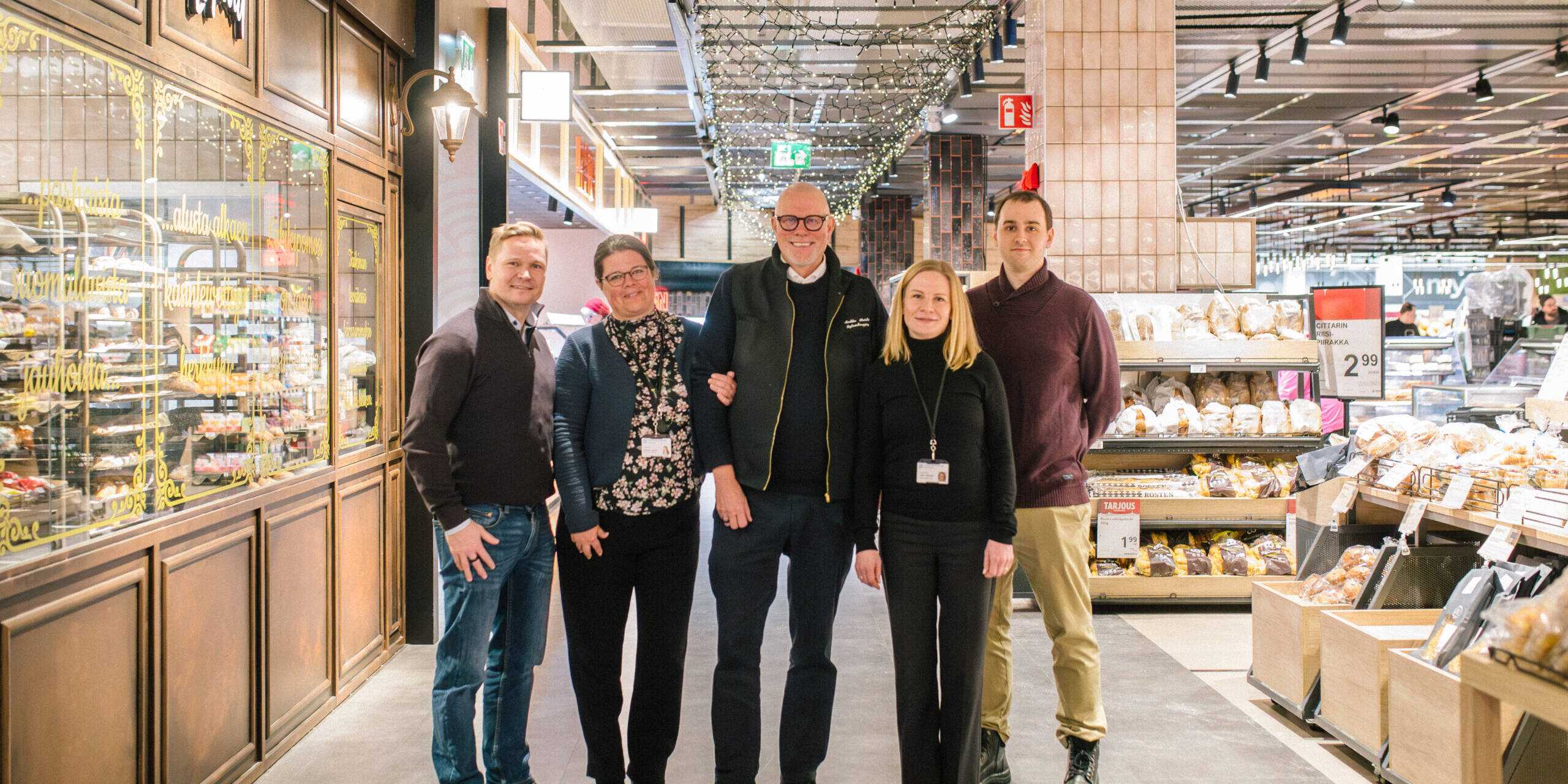Kuvassa viisi henkilöä seisoo hymyillen ruokakaupan käytävällä. Taustalla näkyy leipäosasto ja myymälän sisustusta, jossa on lämminhenkinen valaistus ja koristevaloja katossa. Vasemmalla puolella on tyylikkäästi somistettu lasivitriini, jossa lukee koristeellisella tekstillä suomenkielisiä sanoja. Oikealla puolella näkyy leipähyllyjä, joissa on tarjouksia ja tuotteita esillä. Kaikilla henkilöillä on siistit vaatteet, ja osalla heistä on nimikortit kaulassaan, viitaten mahdollisesti kaupan henkilökuntaan tai yhteistyökumppaneihin.