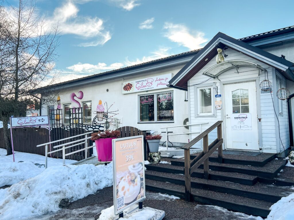 Kakkutalo Gilanin rakennus talvisessa maisemassa. Valkoinen, yksikerroksinen rakennus, jonka seinään on maalattu muraaleja, kuten jäätelötötterö ja koristeellisia kuvioita. Rakennuksen edessä on portaat ja luiska sisäänkäynnille, ja luminen piha on koristeltu vaaleanpunaisilla istutusruukuilla. Edustalla mainoskyltti, jossa mainostetaan uutuustuotetta.