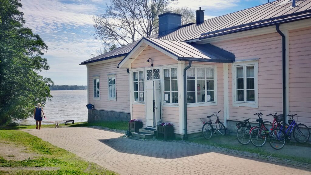 Vaaleanpunainen puutalo Kartanon alueella veden äärellä aurinkoisena päivänä. Etualalla polkupyöriä parkissa ja nainen kävelemässä pienen koiran kanssa kohti järveä.