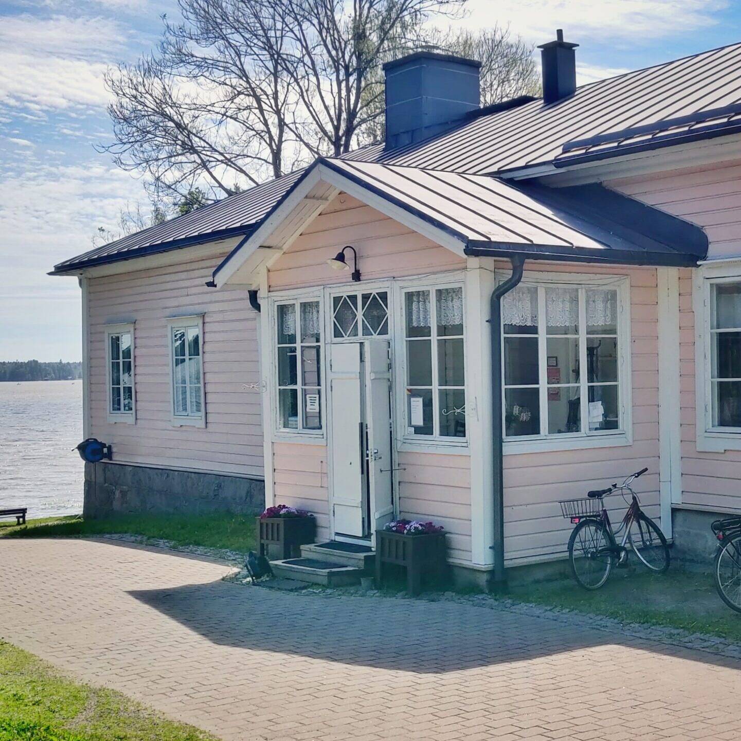 Vaaleanpunainen puutalo Kartanon alueella veden äärellä aurinkoisena päivänä. Etualalla polkupyöriä parkissa ja nainen kävelemässä pienen koiran kanssa kohti järveä.