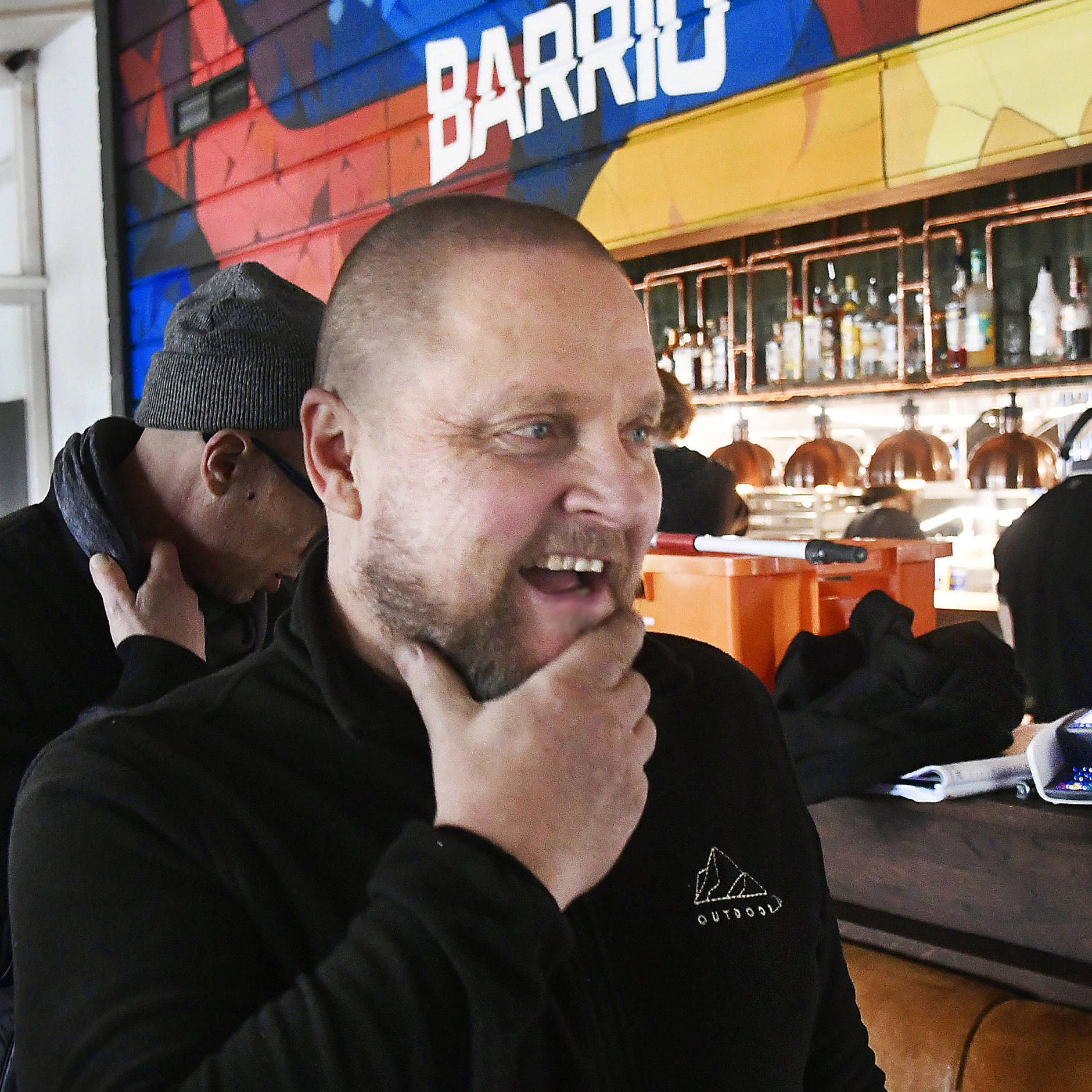 Hymyilevä mies koskettaa leukapartaansa värikkäästi sisustetun baarin edustalla, taustalla näkyy baaritiski, valaisimia ja muita ihmisiä. Seinällä lukee isoilla kirjaimilla 