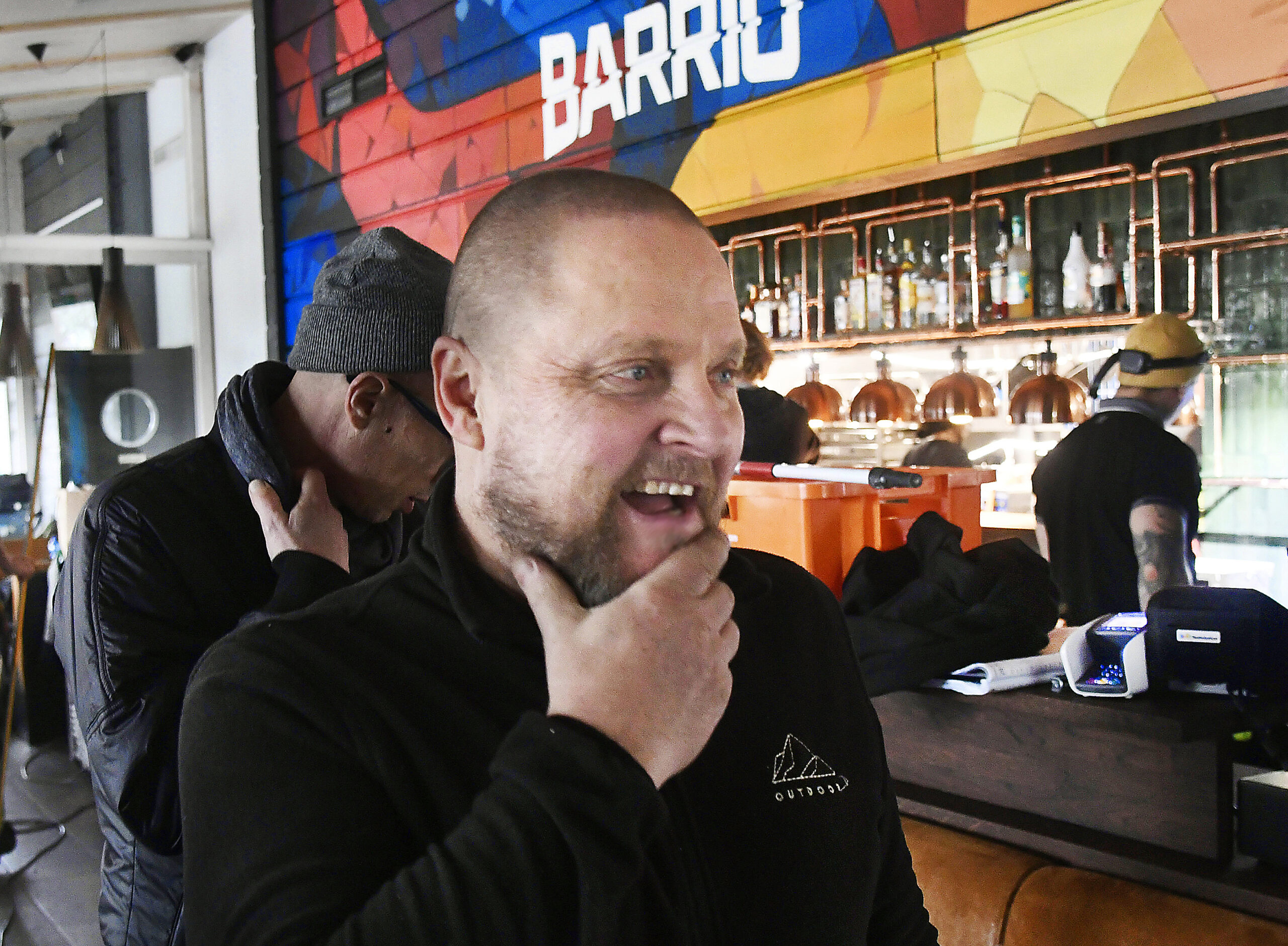 Hymyilevä mies koskettaa leukapartaansa värikkäästi sisustetun baarin edustalla, taustalla näkyy baaritiski, valaisimia ja muita ihmisiä. Seinällä lukee isoilla kirjaimilla "BARRIO".