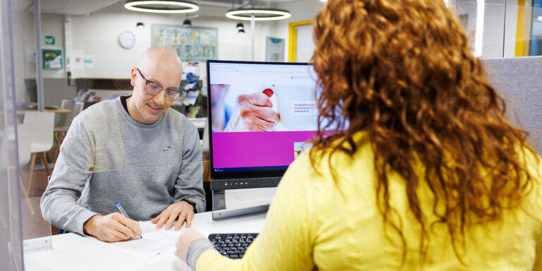 Kalju mies ja kiharatukkainen nainen keskustelevat palvelutiskillä, välissä tietokonenäyttö, jonka ruudulla näkyy käsiä pitelevä kuva ja tekstiä.