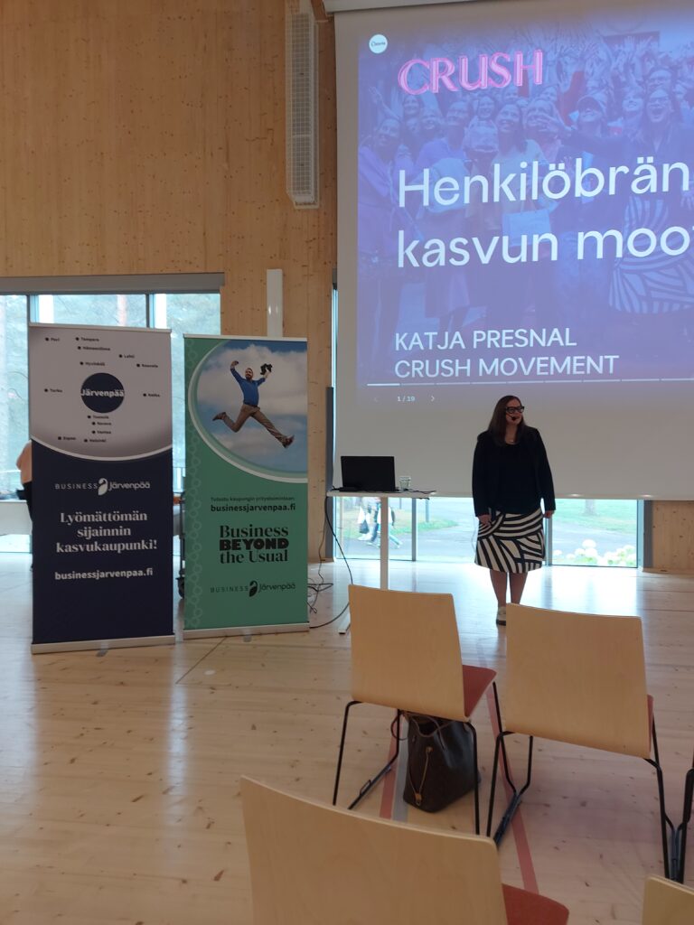 Esitystilanne valoisassa salissa. Etualalla näkyy riveittäin puisia tuoleja, joista osa on tyhjiä. Yhdellä tuolilla on ruskea laukku. Keskellä kuvaa seisoo nainen mustassa jakussa ja mustavalkoraidallisessa hameessa. Hän on esiintymässä suuren valkokankaan edessä, jossa lukee isolla ”CRUSH – Henkilöbrändin kasvun moottori, Katja Presnal, Crush Movement”. Valkokankaan vieressä on kaksi pystybanneria: vasemmalla tummansininen ”Lyömättömän sijainnin kasvukaupunki! Business Järvenpää”, oikealla vihreä ”Business Beyond the Usual”. Taustalla näkyy ikkuna, josta kajastaa päivänvalo.