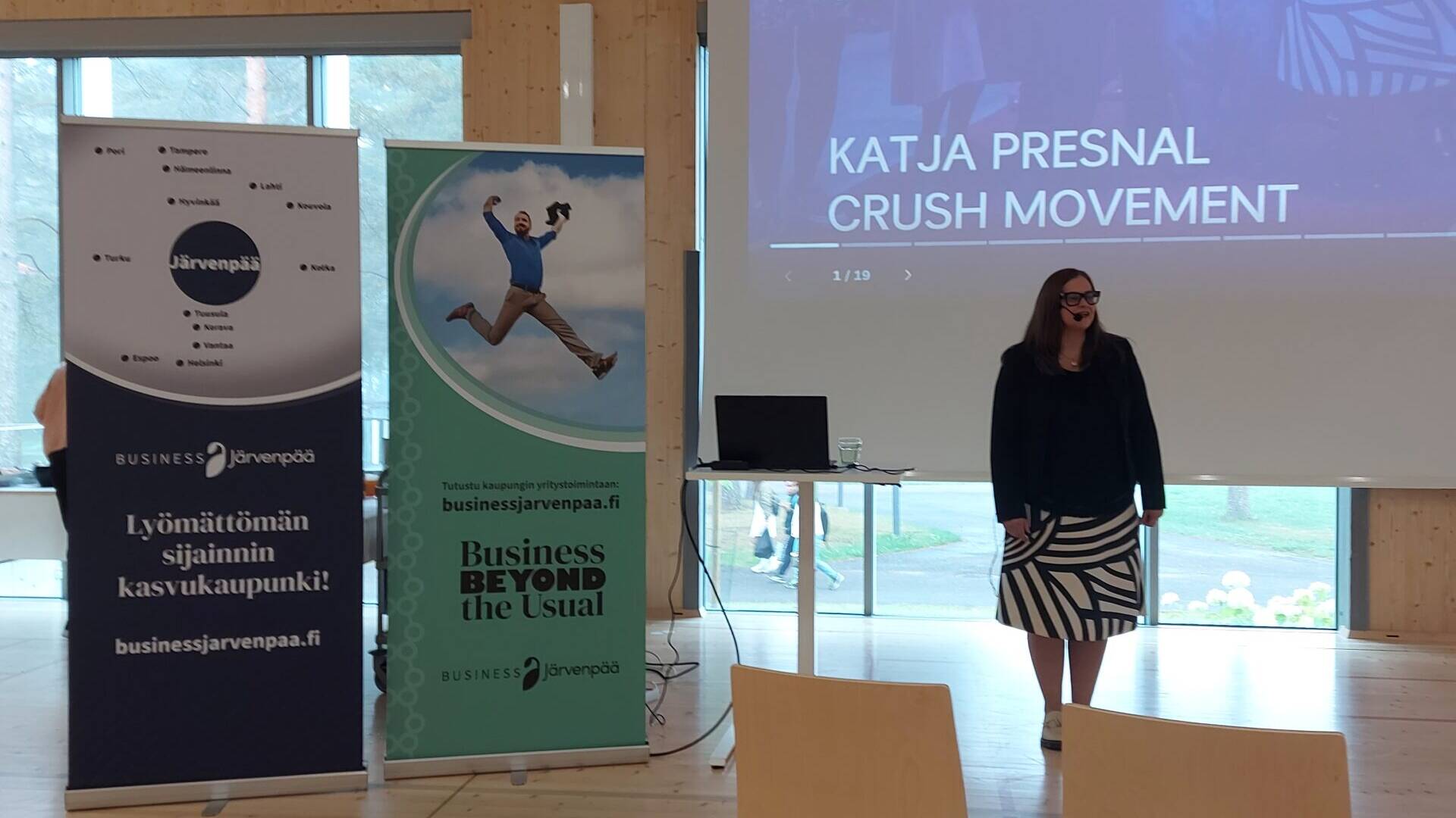 Esitystilanne valoisassa salissa. Etualalla näkyy riveittäin puisia tuoleja, joista osa on tyhjiä. Yhdellä tuolilla on ruskea laukku. Keskellä kuvaa seisoo nainen mustassa jakussa ja mustavalkoraidallisessa hameessa. Hän on esiintymässä suuren valkokankaan edessä, jossa lukee isolla ”CRUSH – Henkilöbrändin kasvun moottori, Katja Presnal, Crush Movement”. Valkokankaan vieressä on kaksi pystybanneria: vasemmalla tummansininen ”Lyömättömän sijainnin kasvukaupunki! Business Järvenpää”, oikealla vihreä ”Business Beyond the Usual”. Taustalla näkyy ikkuna, josta kajastaa päivänvalo.