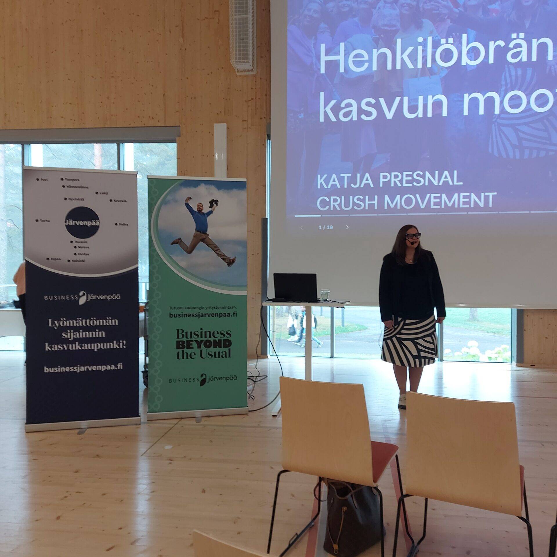 Esitystilanne valoisassa salissa. Etualalla näkyy riveittäin puisia tuoleja, joista osa on tyhjiä. Yhdellä tuolilla on ruskea laukku. Keskellä kuvaa seisoo nainen mustassa jakussa ja mustavalkoraidallisessa hameessa. Hän on esiintymässä suuren valkokankaan edessä, jossa lukee isolla ”CRUSH – Henkilöbrändin kasvun moottori, Katja Presnal, Crush Movement”. Valkokankaan vieressä on kaksi pystybanneria: vasemmalla tummansininen ”Lyömättömän sijainnin kasvukaupunki! Business Järvenpää”, oikealla vihreä ”Business Beyond the Usual”. Taustalla näkyy ikkuna, josta kajastaa päivänvalo.