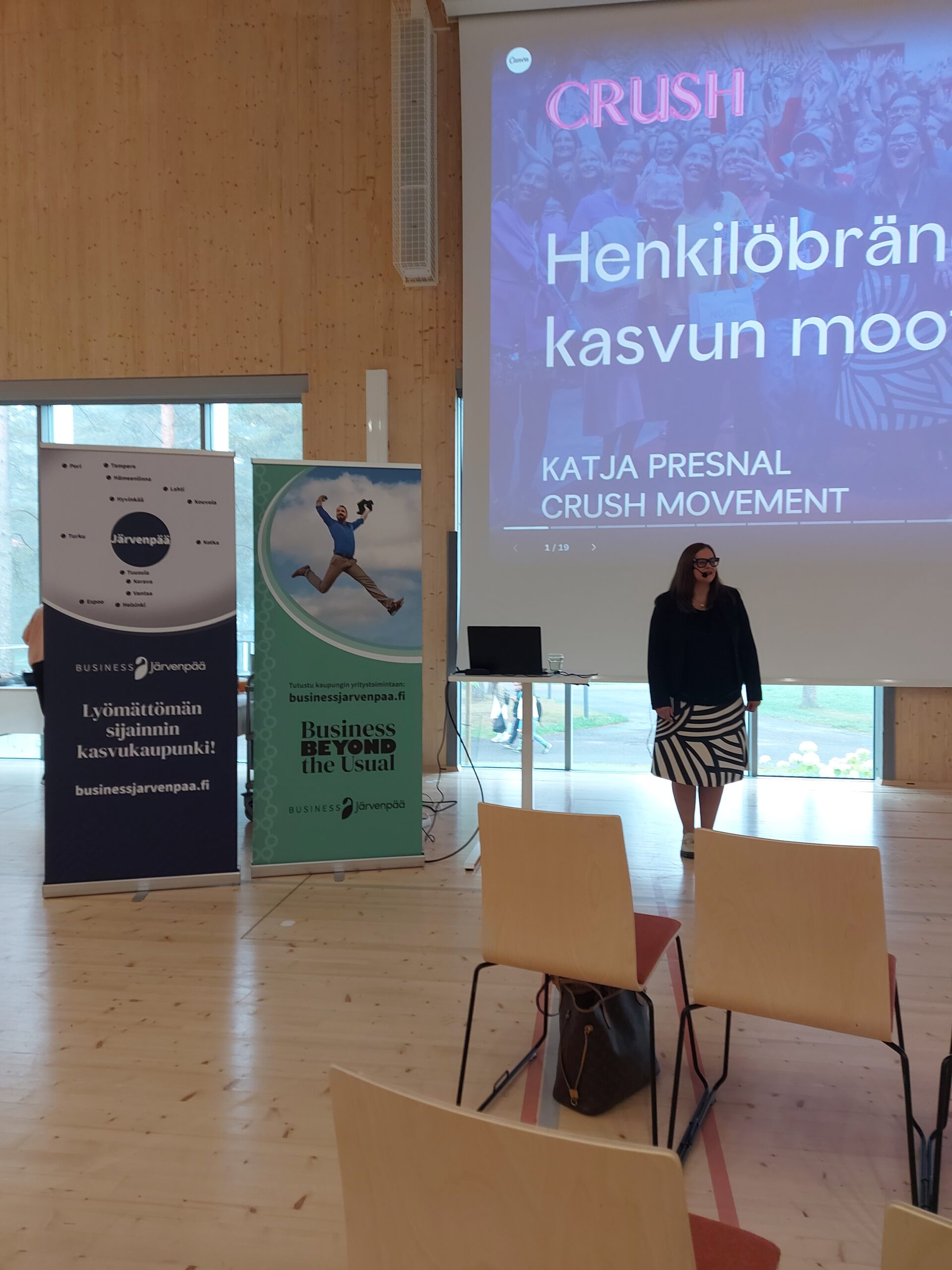 Esitystilanne valoisassa salissa. Etualalla näkyy riveittäin puisia tuoleja, joista osa on tyhjiä. Yhdellä tuolilla on ruskea laukku. Keskellä kuvaa seisoo nainen mustassa jakussa ja mustavalkoraidallisessa hameessa. Hän on esiintymässä suuren valkokankaan edessä, jossa lukee isolla ”CRUSH – Henkilöbrändin kasvun moottori, Katja Presnal, Crush Movement”. Valkokankaan vieressä on kaksi pystybanneria: vasemmalla tummansininen ”Lyömättömän sijainnin kasvukaupunki! Business Järvenpää”, oikealla vihreä ”Business Beyond the Usual”. Taustalla näkyy ikkuna, josta kajastaa päivänvalo.