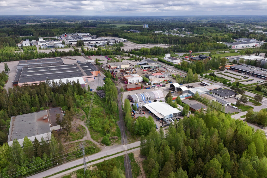 Ilmakuva Järvenpään Jampan teollisuusalueesta ja Wärtsilän tehdasalueesta. Kuvassa näkyy suuria tehdas- ja varastohalleja, pienempiä työpajoja, varastoalueita sekä niiden ympärillä metsää ja viheralueita. Etualalla kulkee kapea junarata, ja taustalla avautuu laaja metsäinen maisema sekä muita teollisuusrakennuksia.