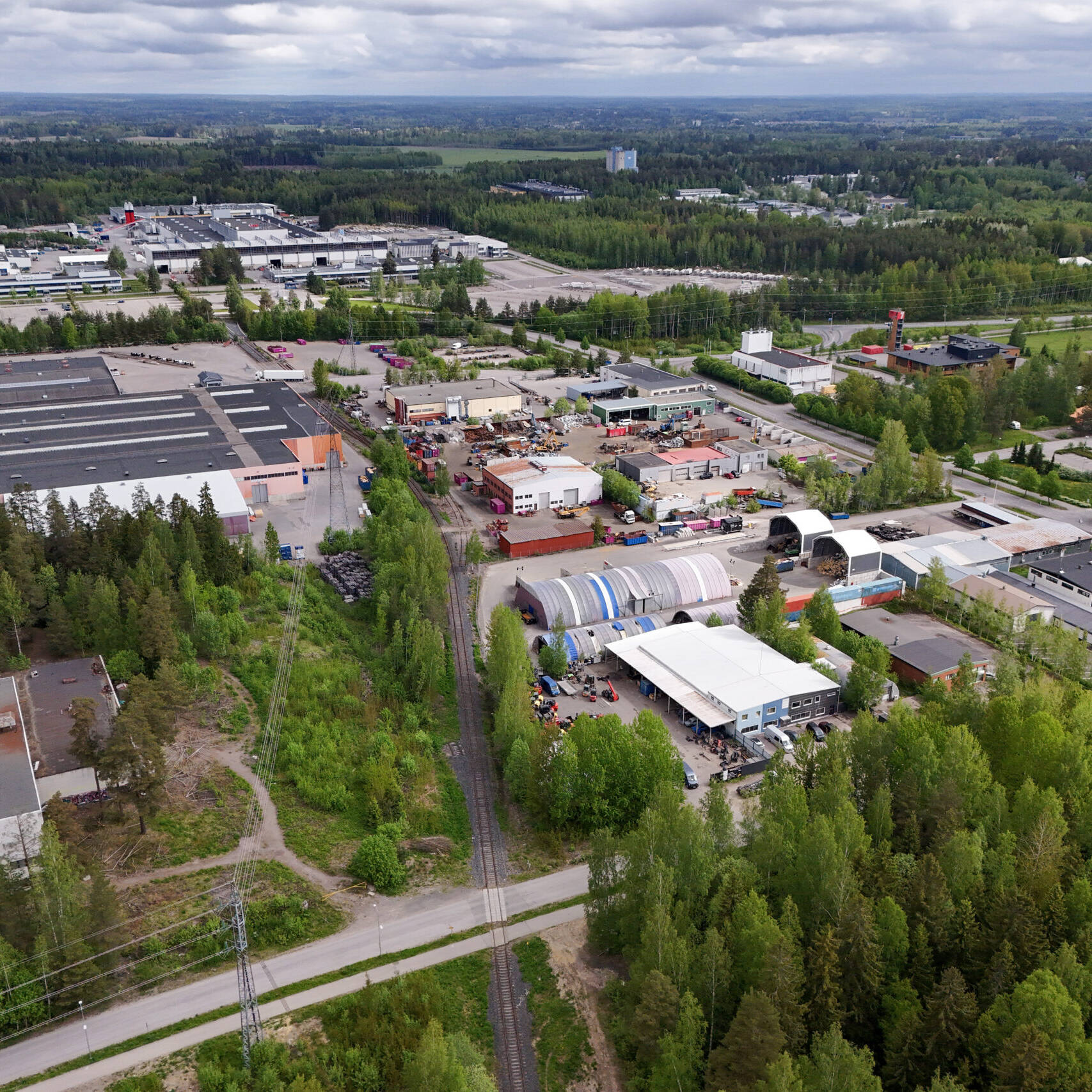 Ilmakuva Järvenpään Jampan teollisuusalueesta ja Wärtsilän tehdasalueesta. Kuvassa näkyy suuria tehdas- ja varastohalleja, pienempiä työpajoja, varastoalueita sekä niiden ympärillä metsää ja viheralueita. Etualalla kulkee kapea junarata, ja taustalla avautuu laaja metsäinen maisema sekä muita teollisuusrakennuksia.