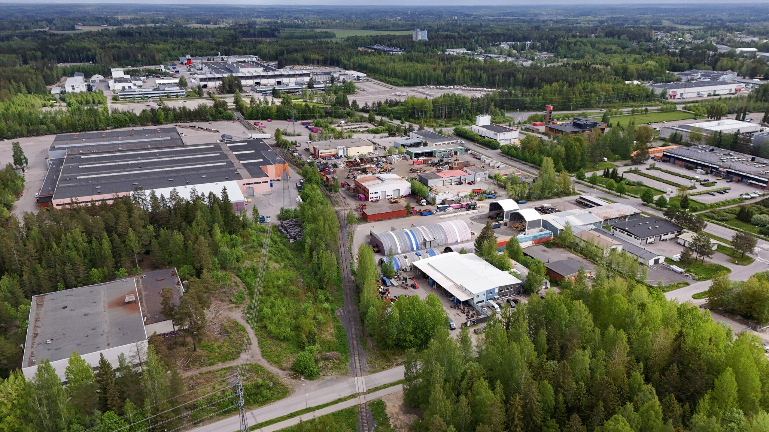 Ilmakuva Järvenpään Jampan teollisuusalueesta ja Wärtsilän tehdasalueesta. Kuvassa näkyy suuria tehdas- ja varastohalleja, pienempiä työpajoja, varastoalueita sekä niiden ympärillä metsää ja viheralueita. Etualalla kulkee kapea junarata, ja taustalla avautuu laaja metsäinen maisema sekä muita teollisuusrakennuksia.