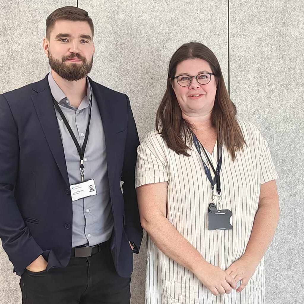 Mies ja nainen seisovat vierekkäin vaalean seinän edessä. Molemmilla on kaulassaan henkilökortit. Mies on puvussa ja nainen raidallisessa mekossa.