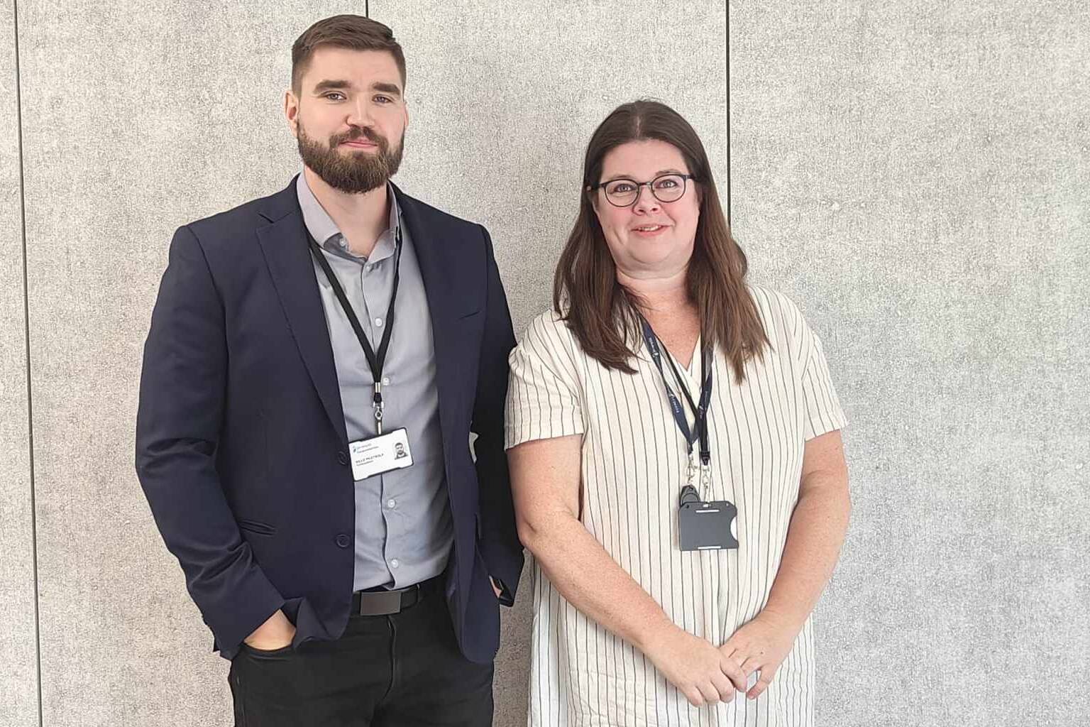 Mies ja nainen seisovat vierekkäin vaalean seinän edessä. Molemmilla on kaulassaan henkilökortit. Mies on puvussa ja nainen raidallisessa mekossa.