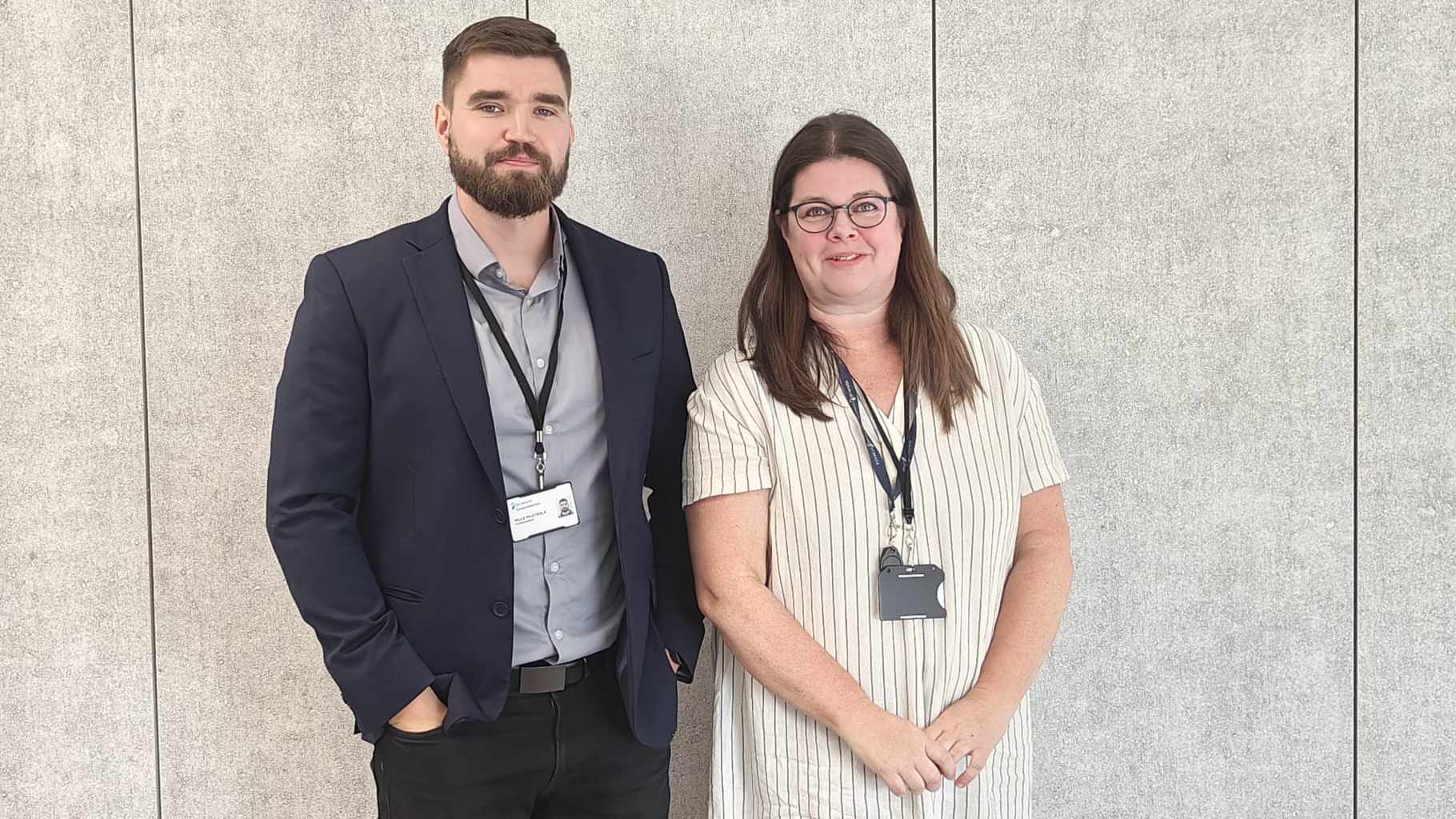 Mies ja nainen seisovat vierekkäin vaalean seinän edessä. Molemmilla on kaulassaan henkilökortit. Mies on puvussa ja nainen raidallisessa mekossa.