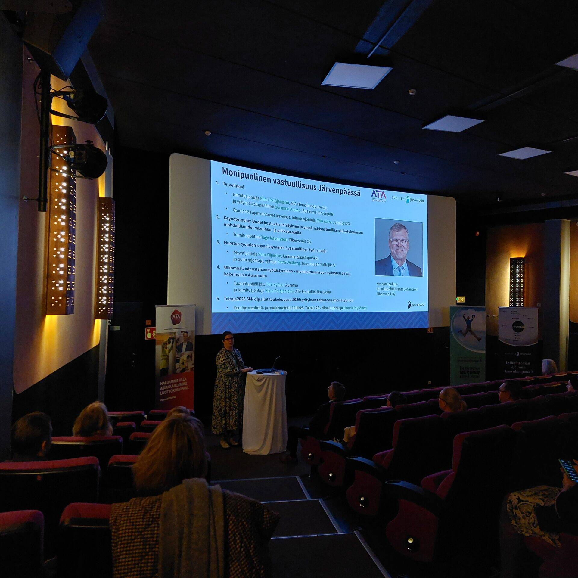 Puhuja seisoo valkokankaan edessä esittelemässä tapahtuman ohjelmaa nimeltä ”Monipuolinen vastuullisuus Järvenpäässä”. Näytöllä näkyy luettelo esityksistä ja puhujista, ja yleisö seuraa tilaisuutta elokuvateatterin istuimilta.