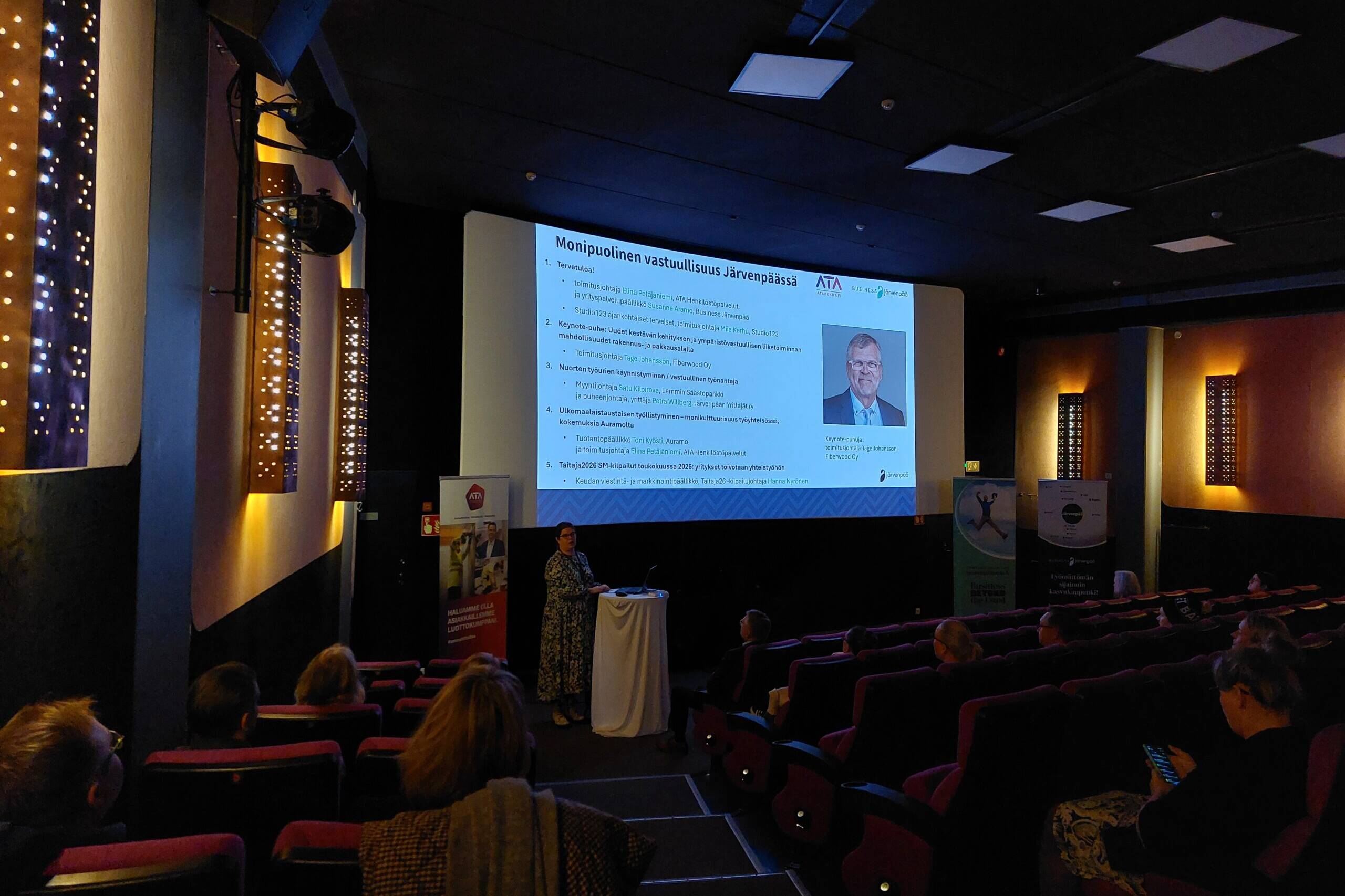 Puhuja seisoo valkokankaan edessä esittelemässä tapahtuman ohjelmaa nimeltä ”Monipuolinen vastuullisuus Järvenpäässä”. Näytöllä näkyy luettelo esityksistä ja puhujista, ja yleisö seuraa tilaisuutta elokuvateatterin istuimilta.