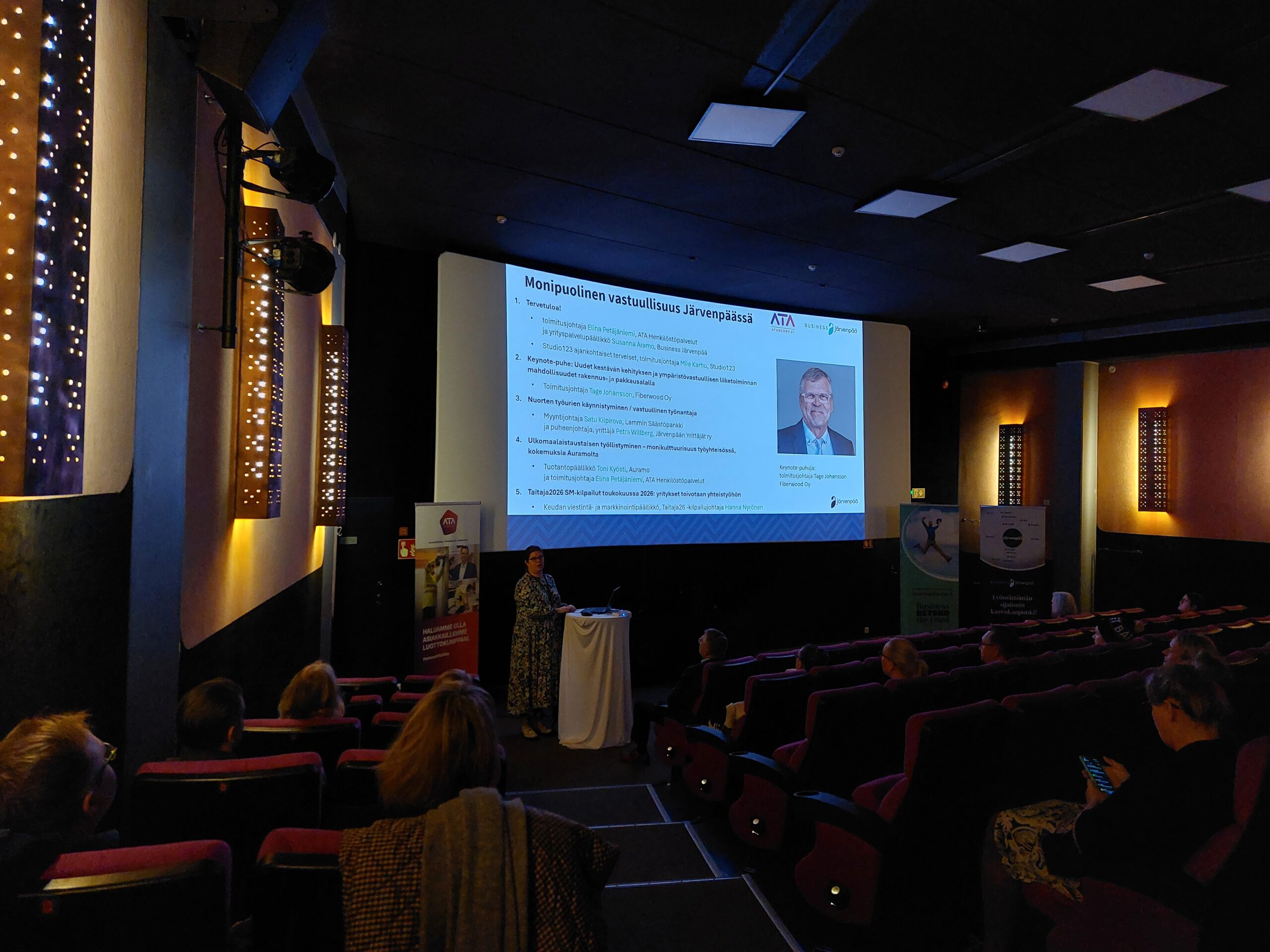 Puhuja seisoo valkokankaan edessä esittelemässä tapahtuman ohjelmaa nimeltä ”Monipuolinen vastuullisuus Järvenpäässä”. Näytöllä näkyy luettelo esityksistä ja puhujista, ja yleisö seuraa tilaisuutta elokuvateatterin istuimilta.