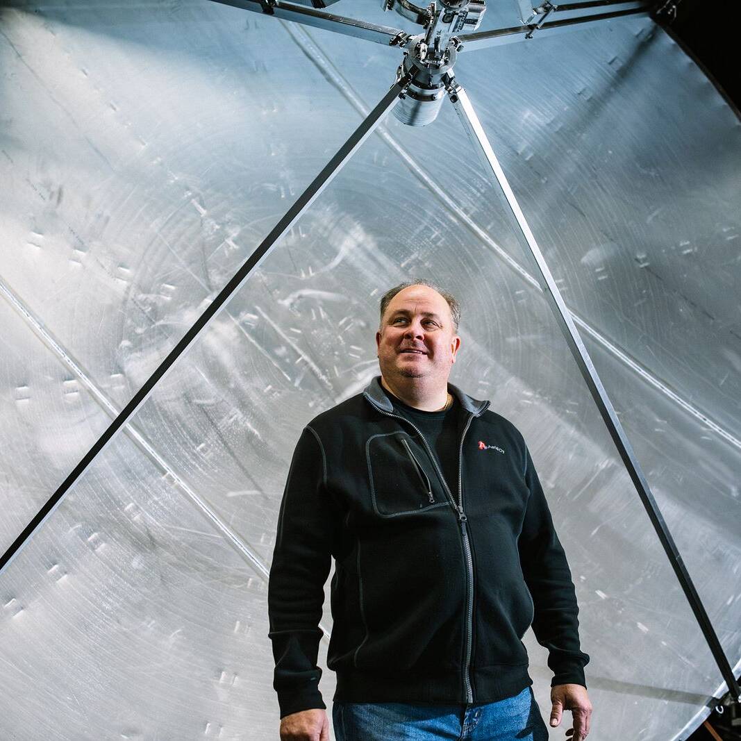 Mies seisoo suuren pyöreän metallisen rakenteen edessä sisätiloissa. Taustalla näkyy suuri kiiltävä metallipinta, johon on kiinnitetty tukirakenteita. Mies hymyilee hieman ja katsoo sivulle.