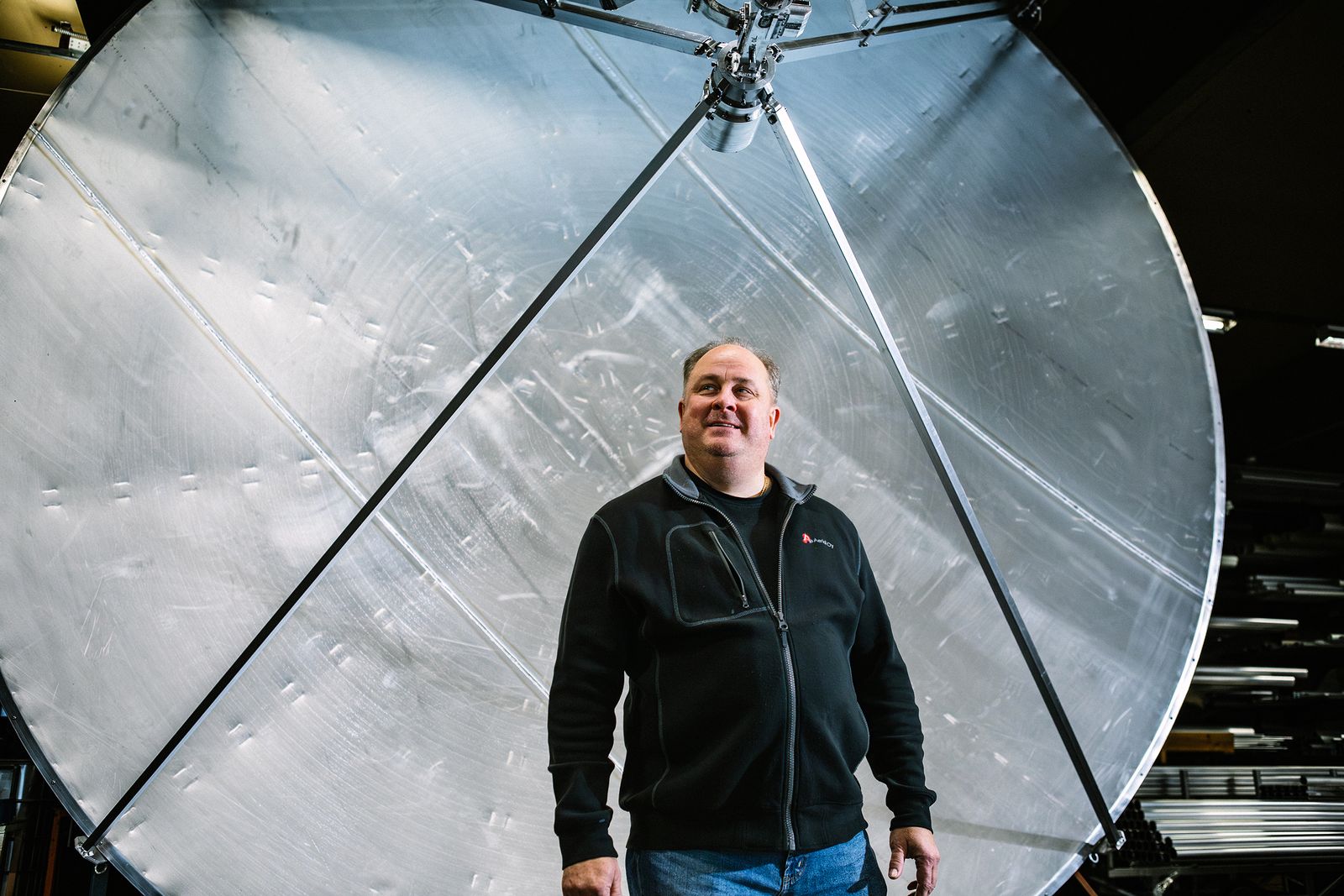 Mies seisoo suuren pyöreän metallisen rakenteen edessä sisätiloissa. Taustalla näkyy suuri kiiltävä metallipinta, johon on kiinnitetty tukirakenteita. Mies hymyilee hieman ja katsoo sivulle.